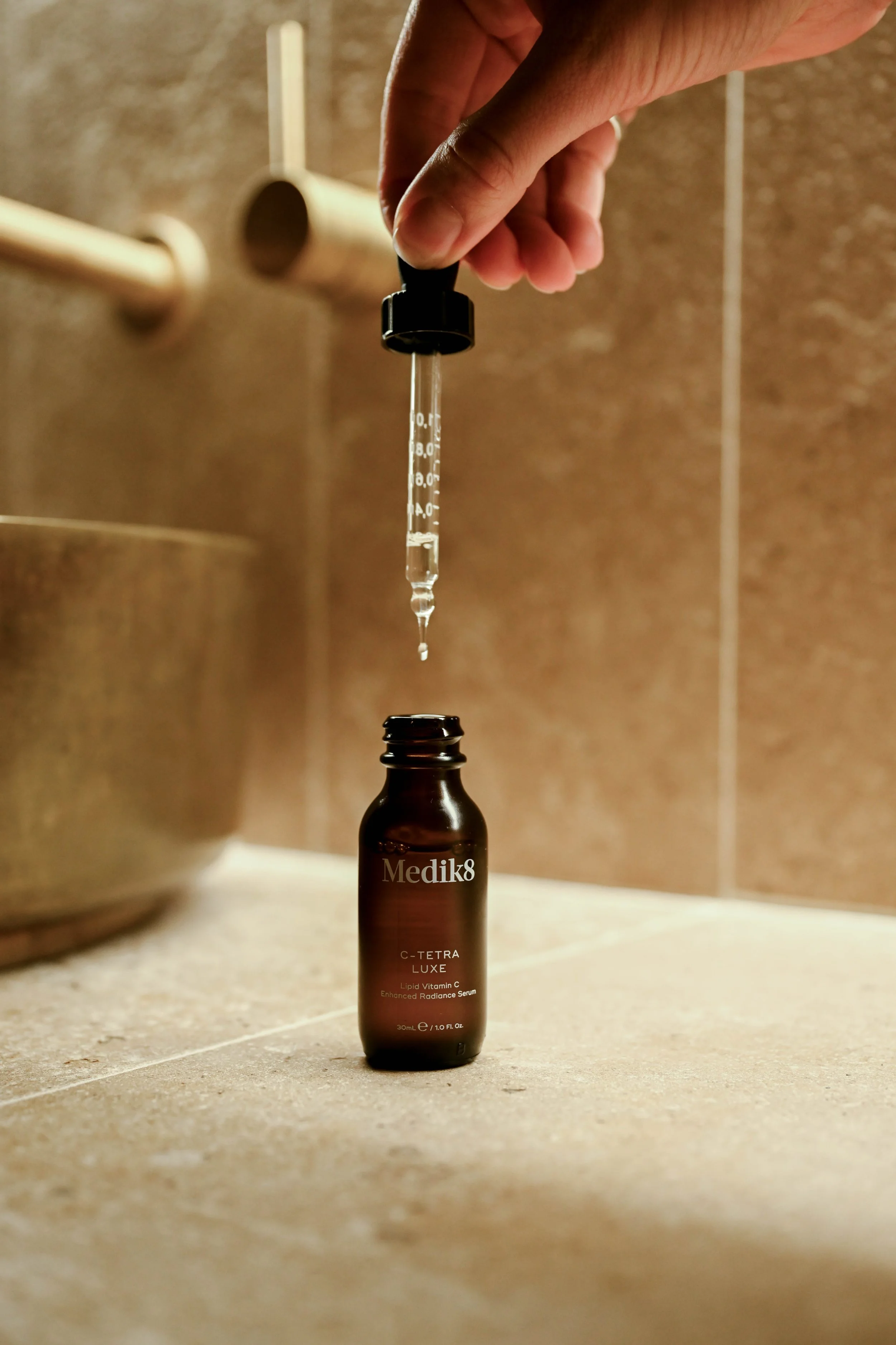 A dropper with a black top is held above a small amber bottle labeled "Medik8 C-Tetra Luxe Lipid Vitamin C Enhanced Radiance Serum." The background has beige tiled walls, and the setting appears to be a bathroom or skincare area.