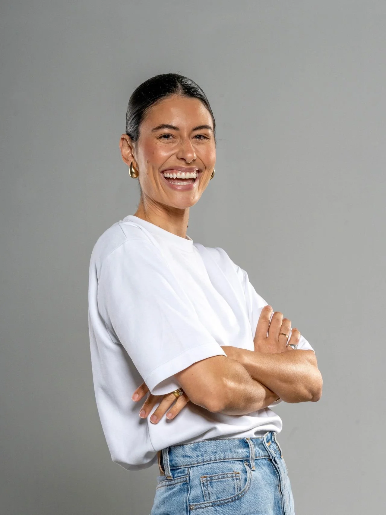 A woman smiling with arms crossed, wearing a white t-shirt and blue jeans against a plain gray background.