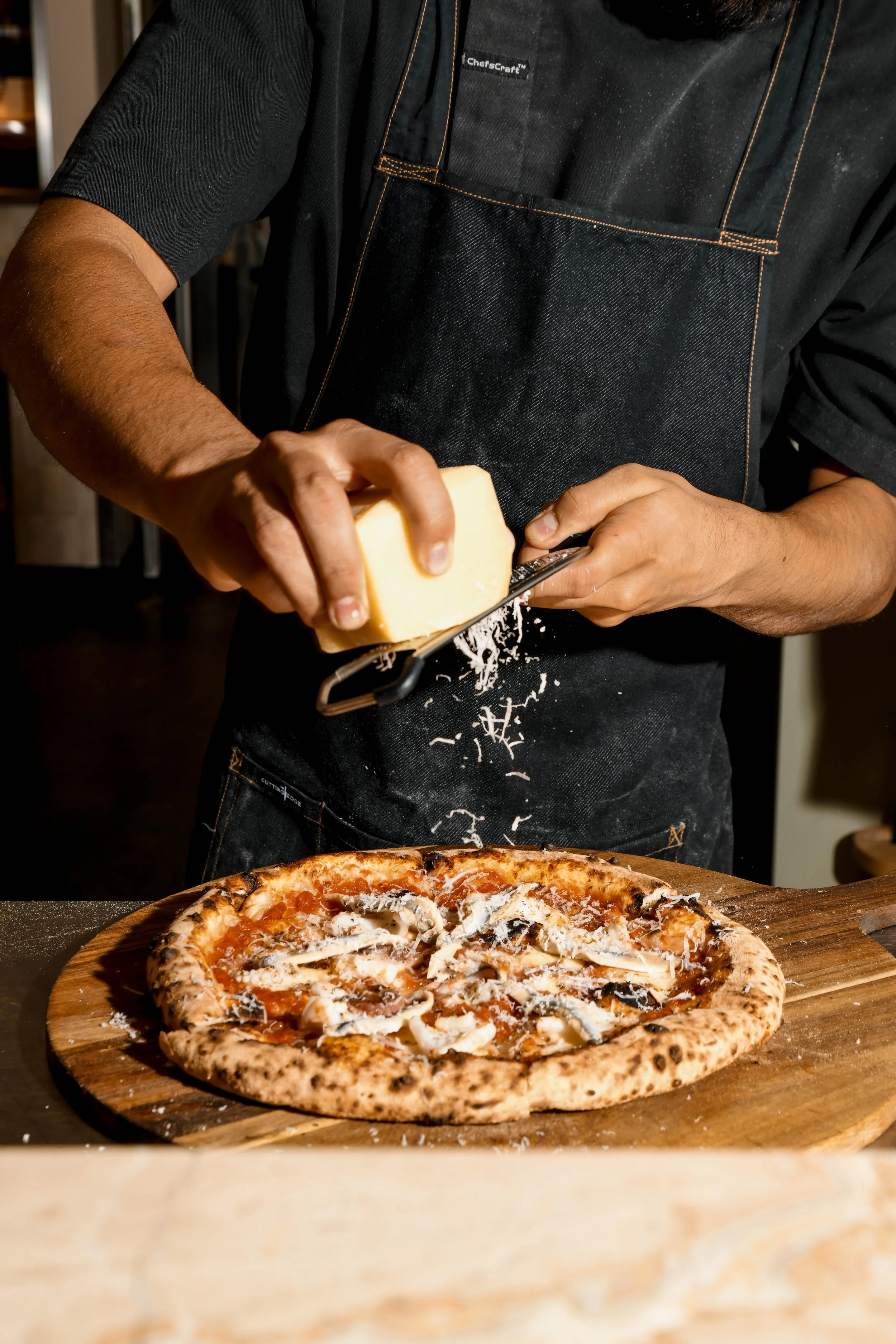 Person grating cheese over a pizza on a wooden pizza peel, wearing a black apron and a black short-sleeved shirt.