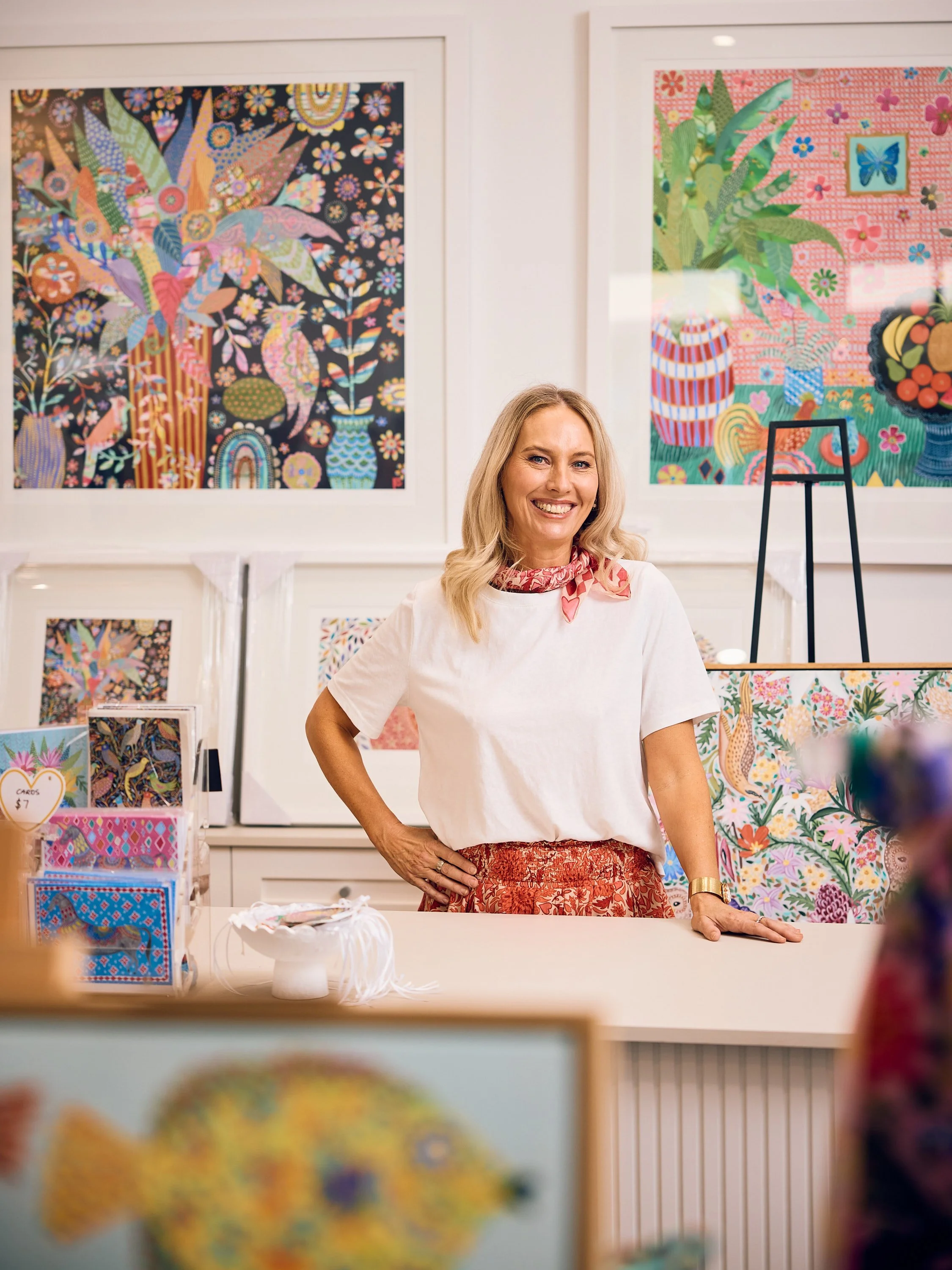 A woman standing behind a counter at an art gallery, smiling, surrounded by colorful abstract artwork on the walls, with craft items for sale on the counter.