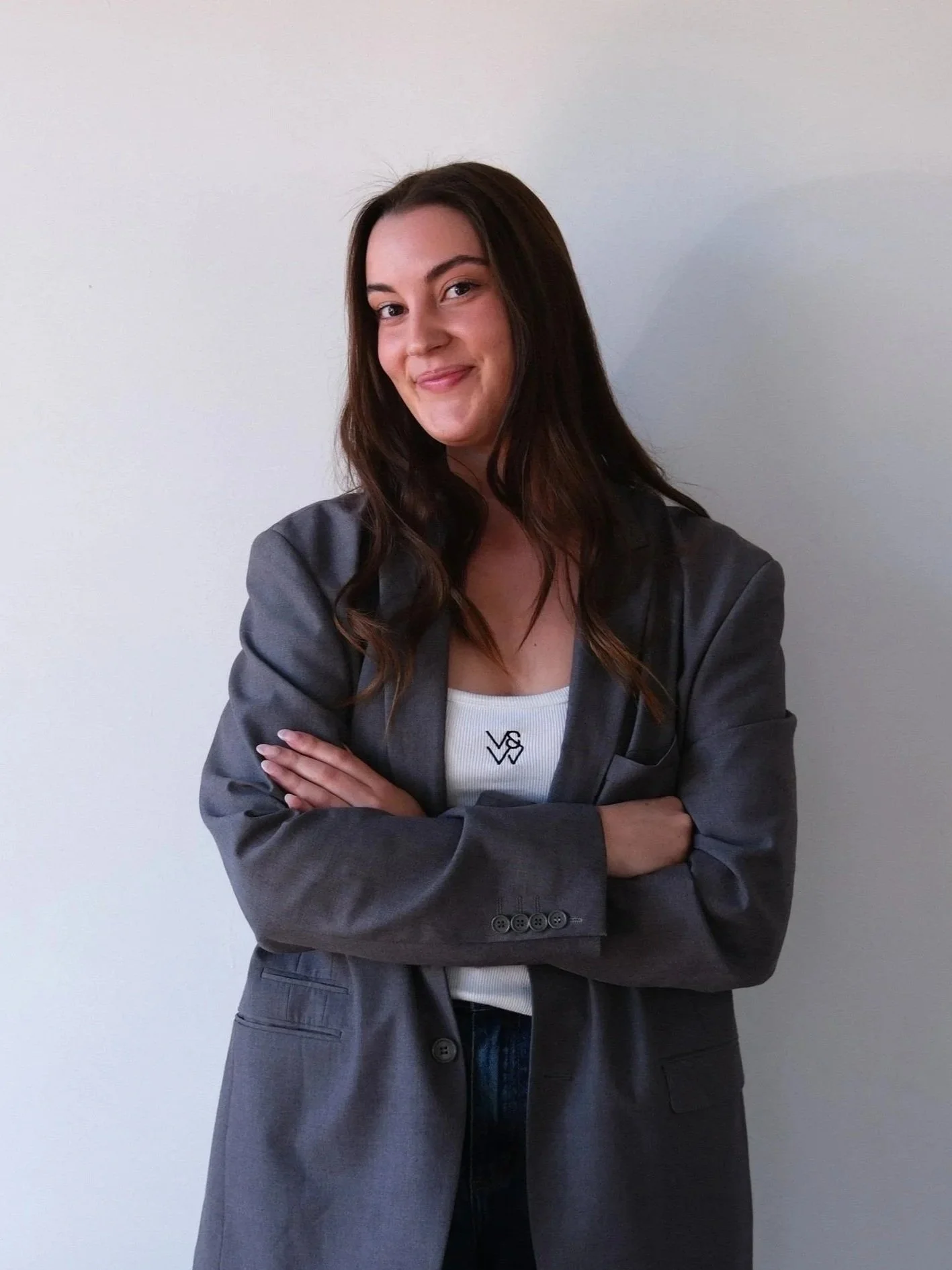 A woman with long brown hair, in a gray blazer and white top, standing with her arms crossed against a plain white wall.