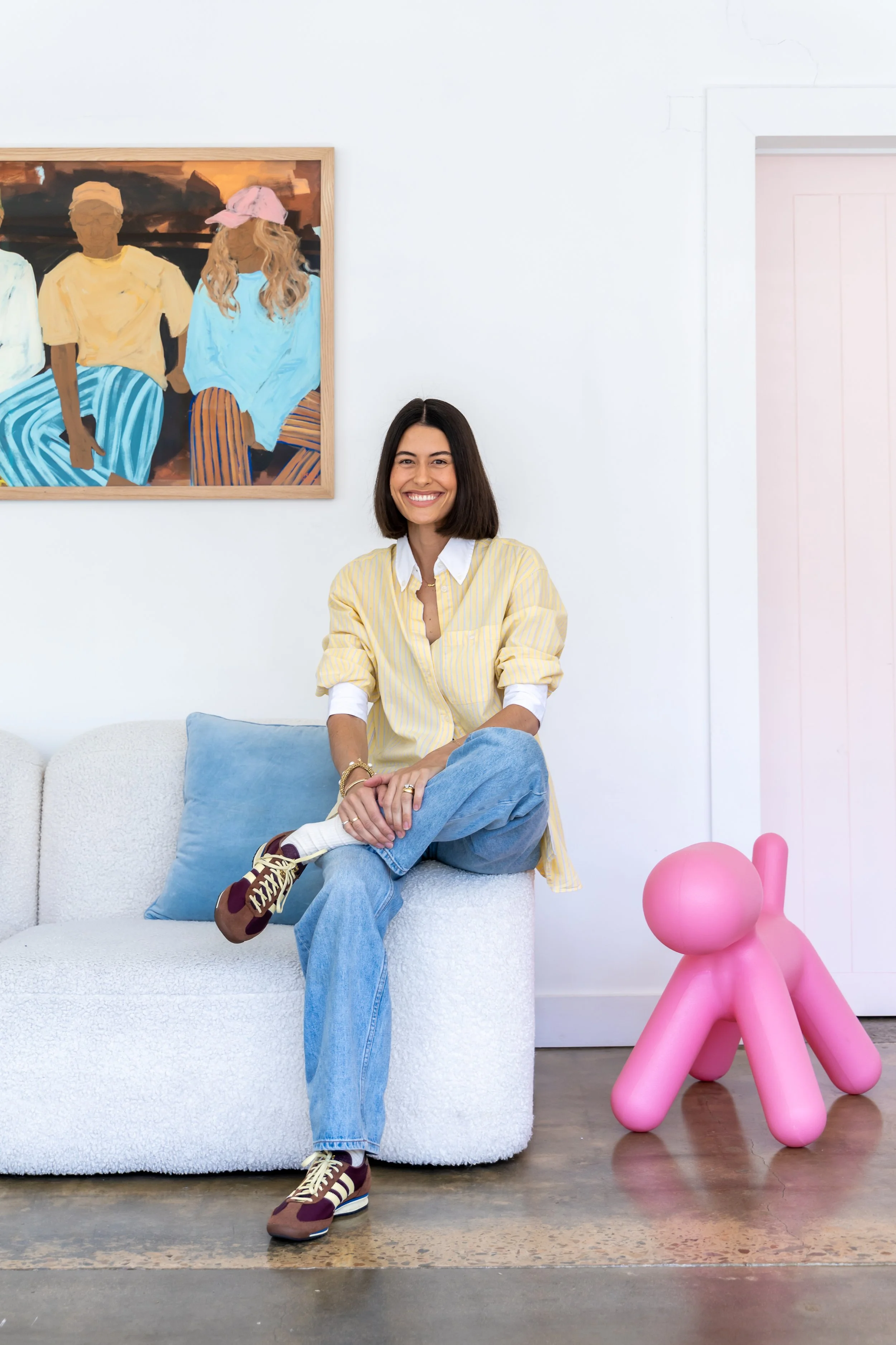 A woman with shoulder-length dark hair sitting on a white textured sofa with a blue pillow, wearing a yellow and white striped shirt, blue jeans, and colorful sneakers, smiling at the camera. Behind her is a large painting of three people sitting, one of whom is wearing a pink hat, on a wall. To her right on the floor is a pink dog-shaped sculpture.