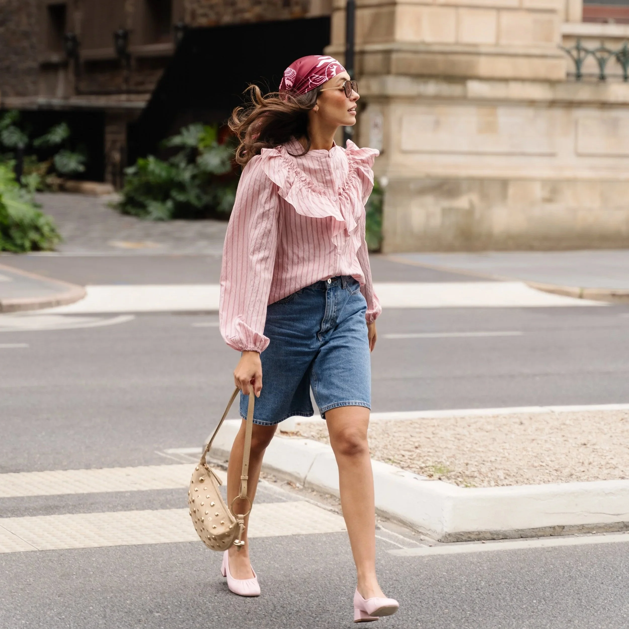 A woman walking outdoors wearing sunglasses, a pink headscarf, a pink ruffled blouse, blue denim shorts, pink high heels, and carrying a cream-colored purse with studs.
