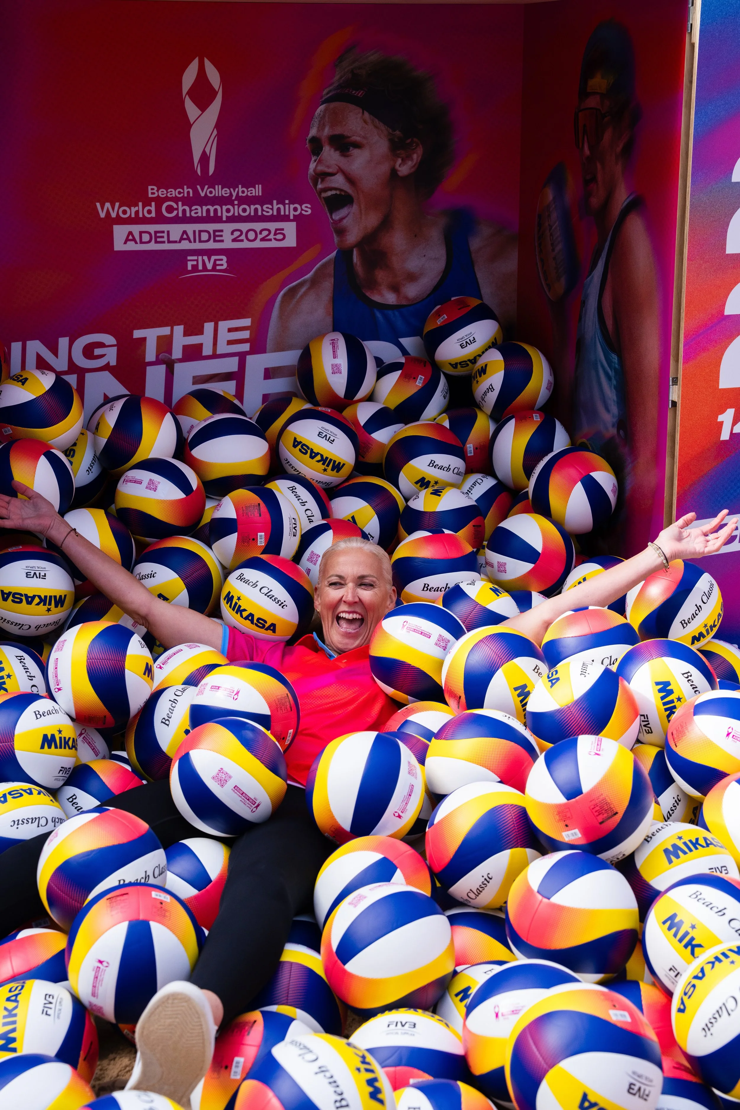 Women in a pink shirt joyfully lying on a pile of Mikasa beach volleyballs at the 2025 Beach Volleyball World Championships in Adelaide, with a large promotional poster behind featuring a female athlete and event details.