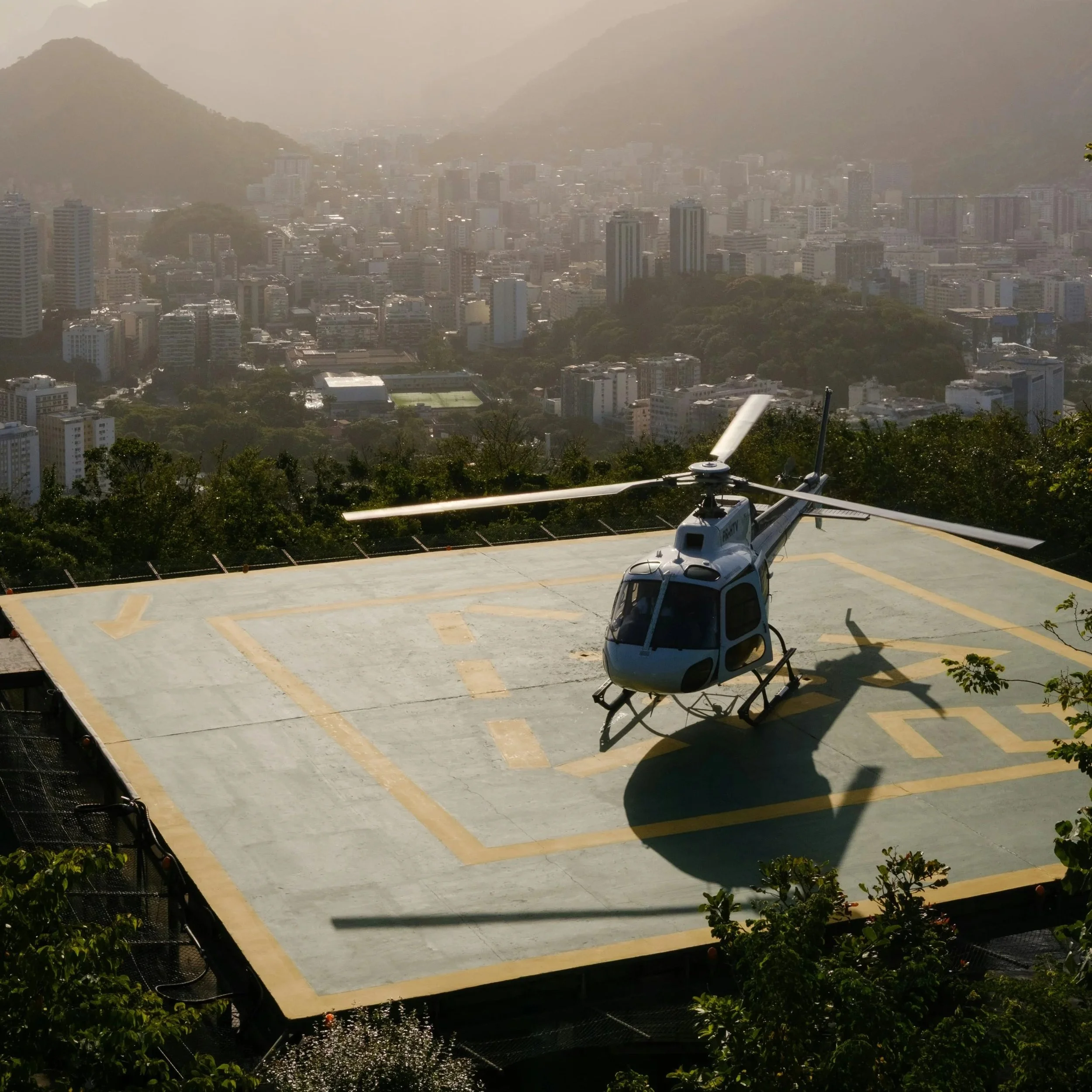A city skyline with tall buildings and mountains in the background under a hazy sky. In the foreground, there is a rooftop with a helicopter landing on helipad.