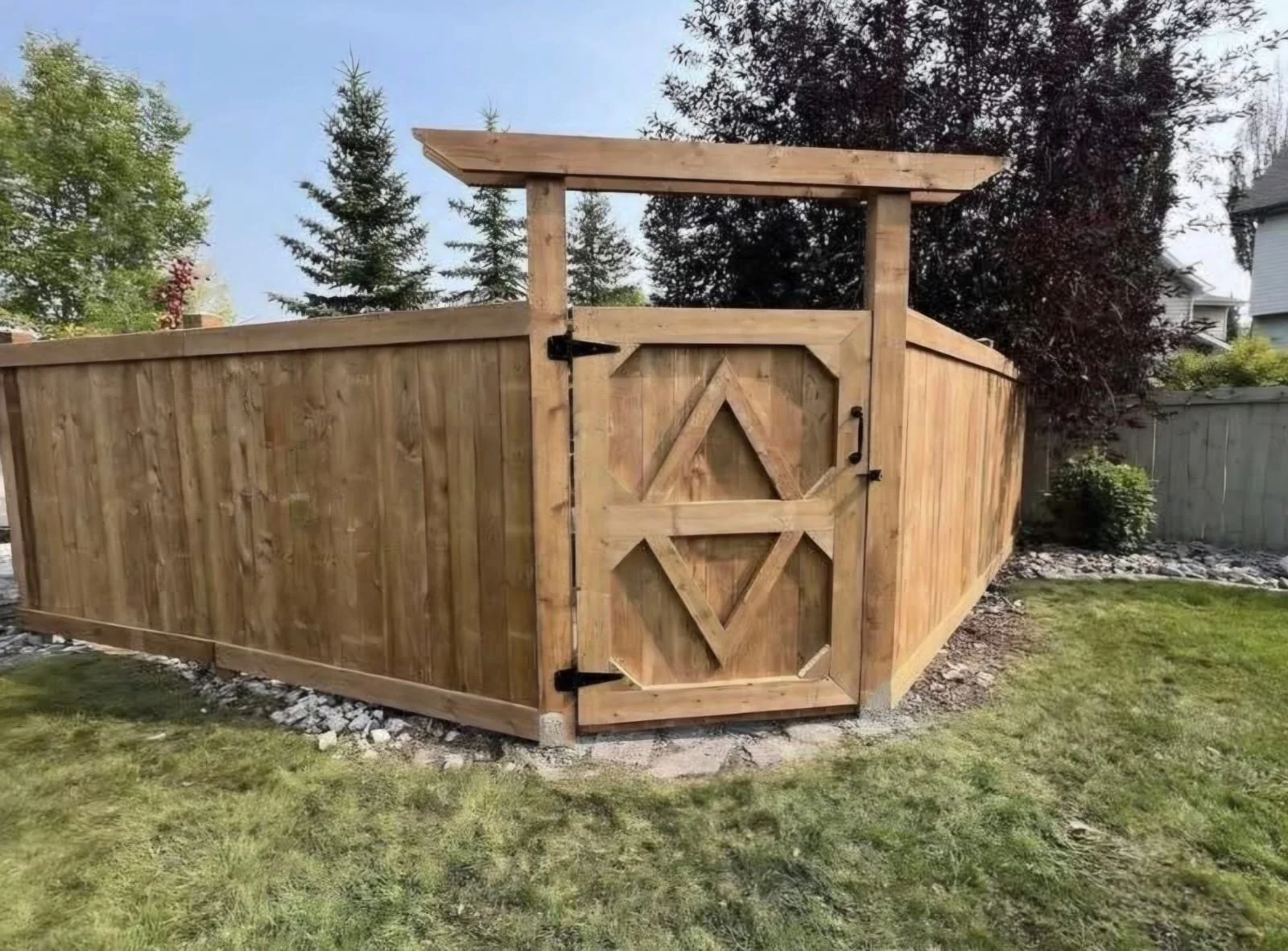 A newly built wooden fence with a decorative gate, surrounding a yard with green grass, trees, and shrubbery.