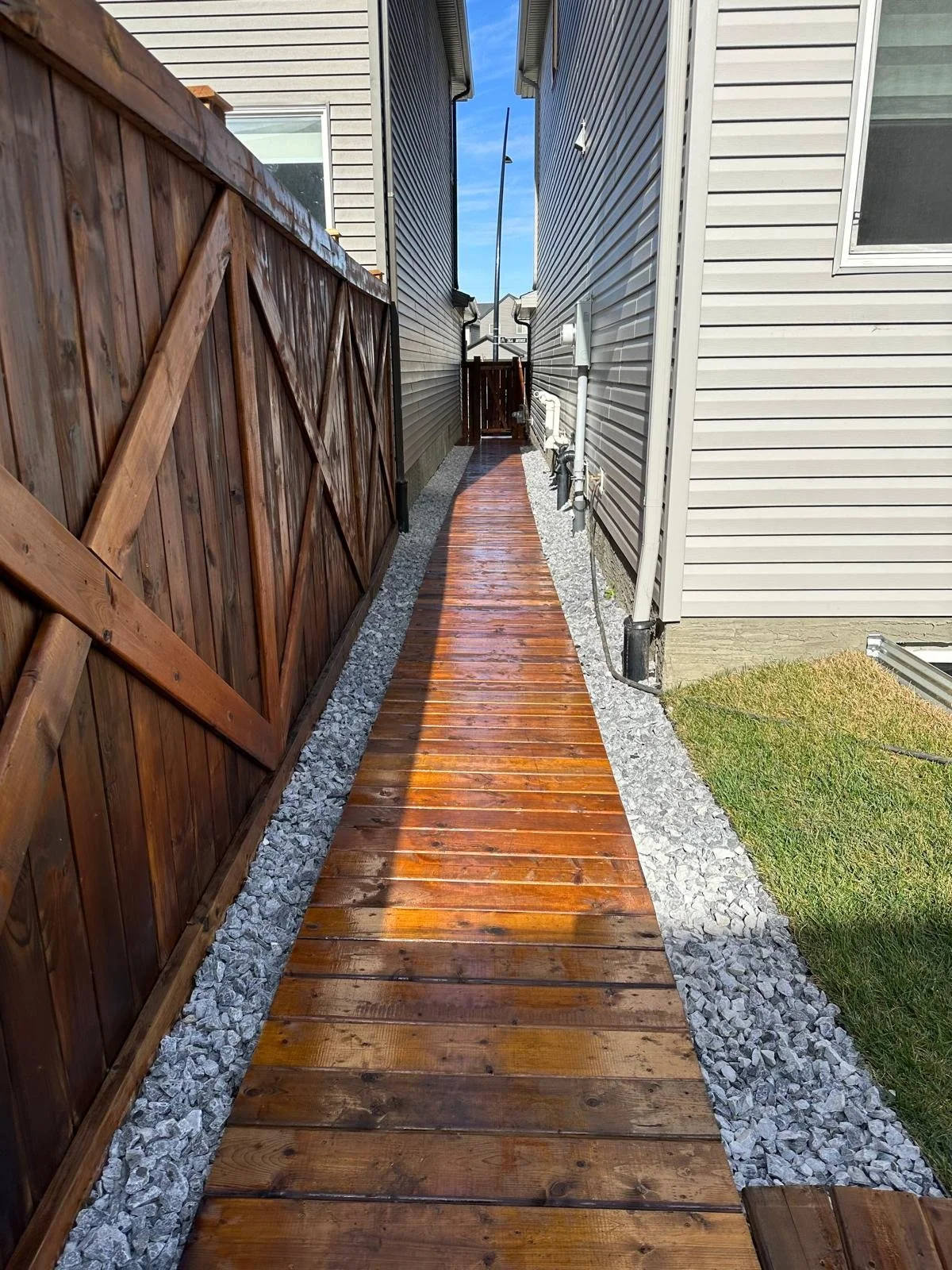 A narrow wooden walkway between two houses, with gravel on either side and a wooden fence on the left.