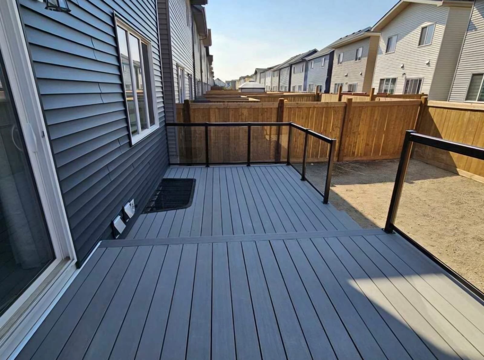 A small outdoor balcony or patio area with a wooden deck, black metal railing, gray siding house, and a neighboring row of similar houses with fenced yards.