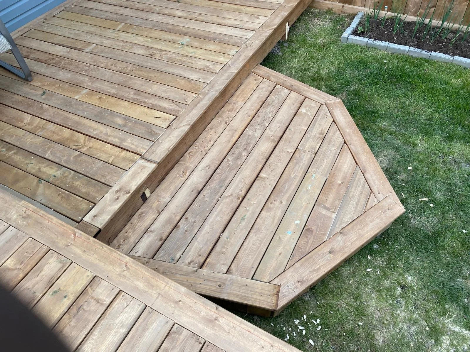 Custom-built multi-level wooden deck with a raised platform and angled design, offering a stylish and functional outdoor living space bordered by a well-maintained lawn and landscaped garden.