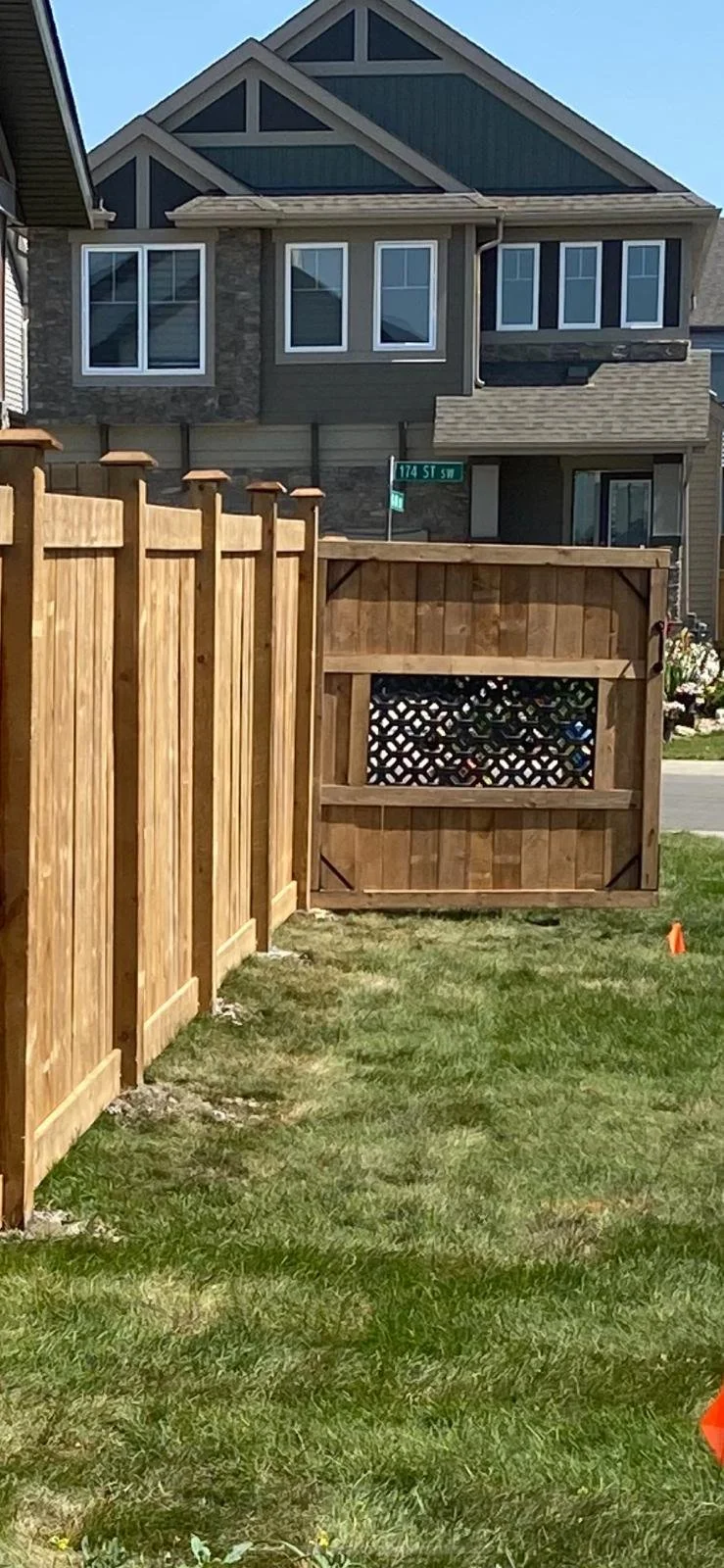 New wooden backyard fence with lattice closing a gate, green grass, and a large modern house with multiple windows in the background.