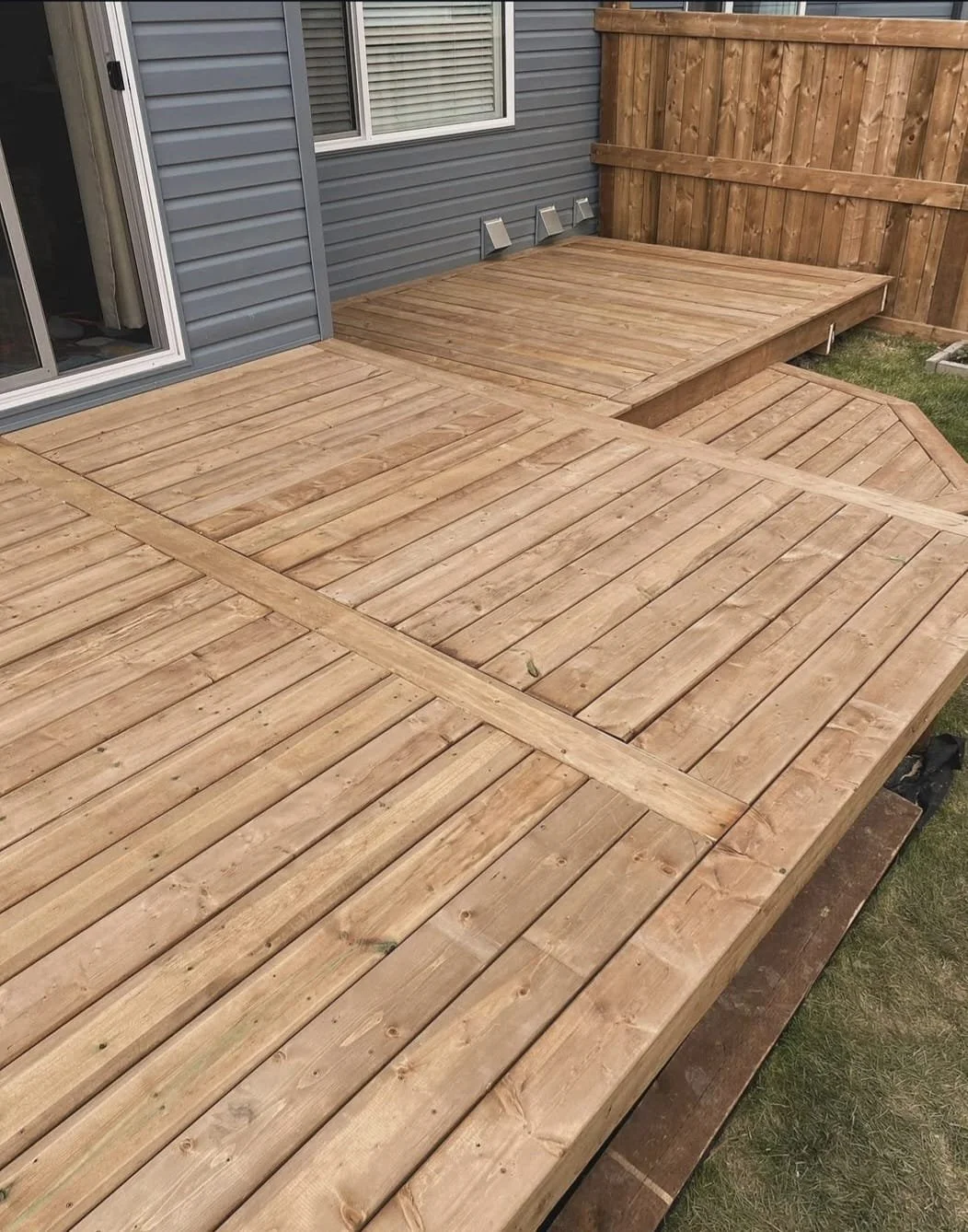 Newly built wooden deck with multiple levels, attached to a house with gray siding, and enclosed by a wooden privacy fence.