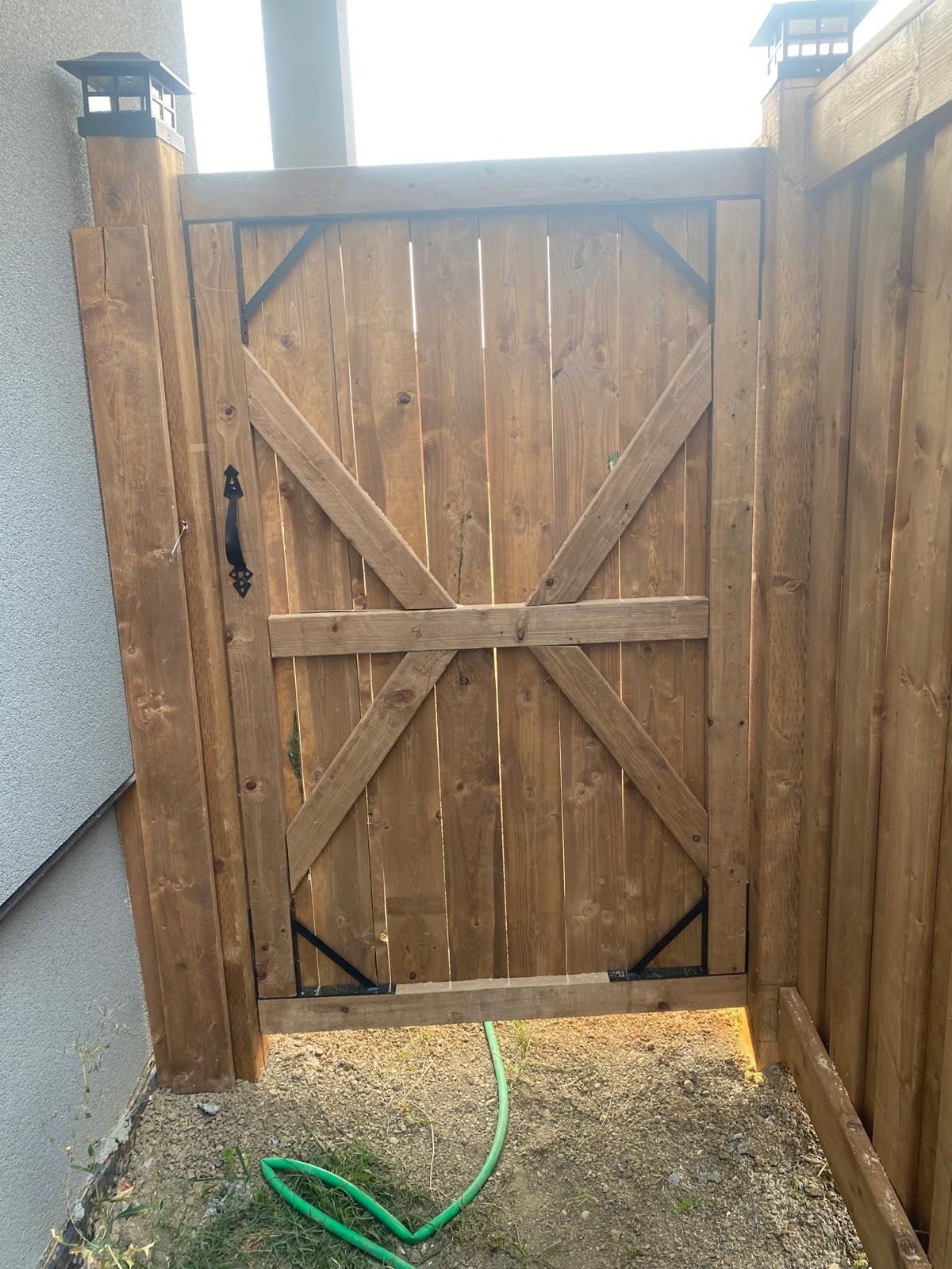 Wooden backyard gate with black hardware, installed between wooden fence posts, with a garden sprinkler hose and grass below.