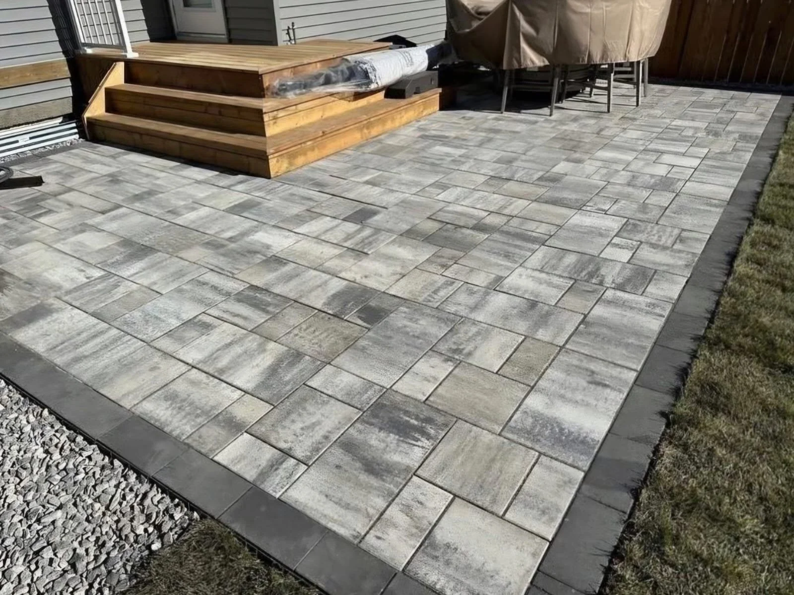 A newly paved backyard patio with stone tiles and a small wooden deck with stairs, surrounded by a border of darker square pavers, and a grassy area along the edge.