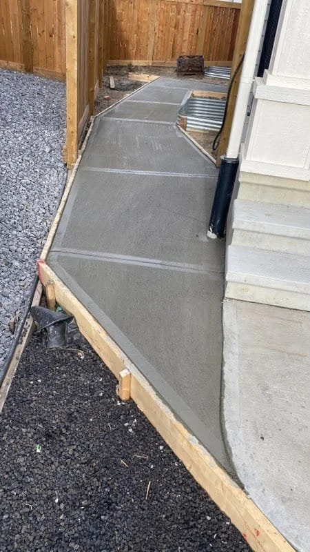 Concrete walkway under construction next to house with wooden and gravel areas.