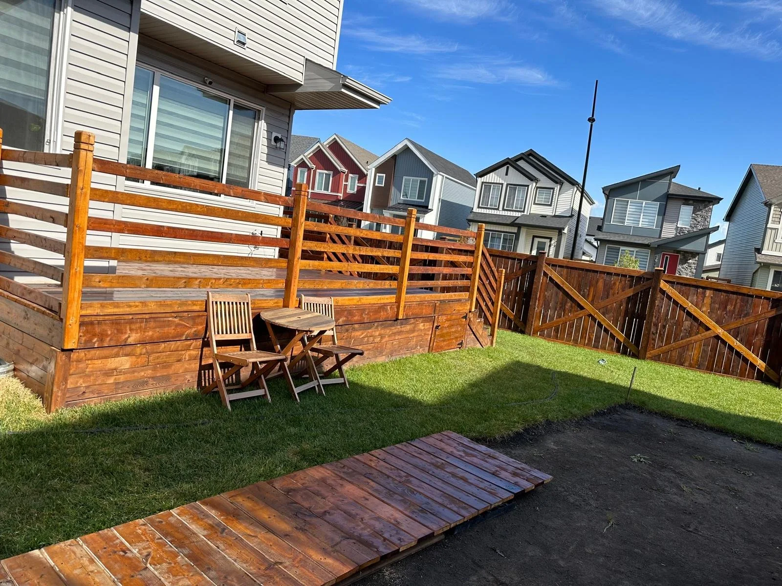 A backyard with a newly built wooden deck, a small wooden table with two chairs, and a grassy lawn enclosed by a wooden fence. Modern multi-story houses are visible in the background under a blue sky.