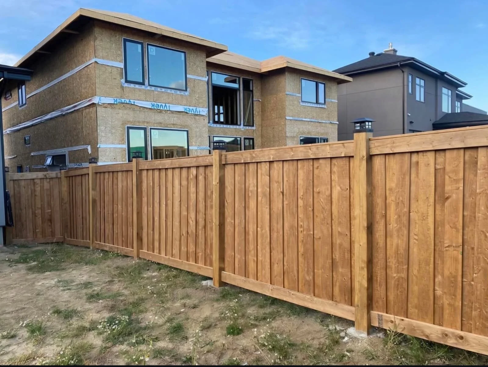 Newly constructed two-story house behind a wooden fence, with visible windows and some finishing work in progress, under a blue sky.