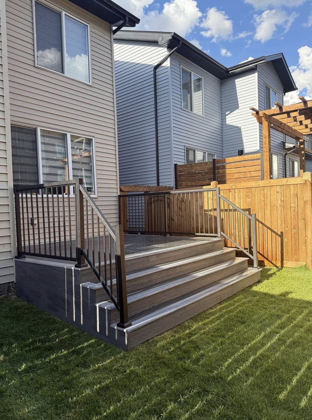 A new outdoor deck with steps and black metal railings attached to a beige house with vinyl siding. The deck has a wooden floor, and there is a wooden privacy fence around part of the yard. The sky is partly cloudy with blue sky visible.