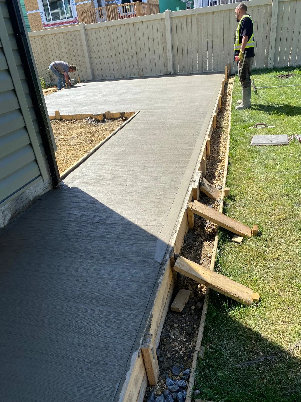 Construction workers are pouring and smoothing concrete for a new sidewalk or patio, with one worker near the house and another on the far side of the frame, with a wooden form and tools visible.