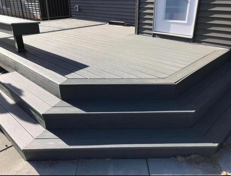A modern wooden deck with gray plank flooring, attached to a house and designed with stairs leading down to a lower level. Finished with black railings for a clean, contemporary look.