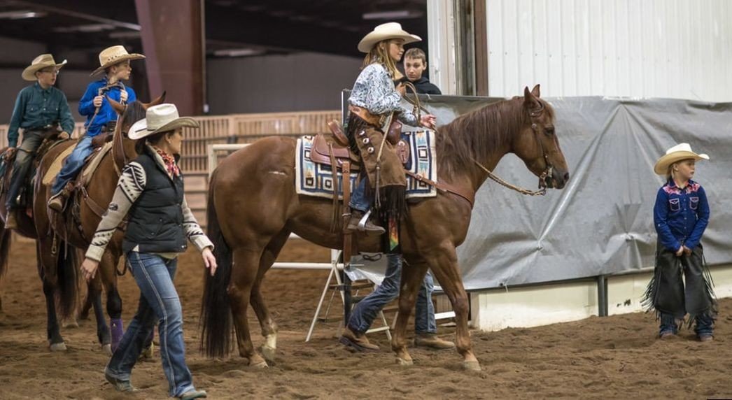 Children riding horses with adults guiding them inside an indoor arena, wearing cowboy hats and Western-style clothing.