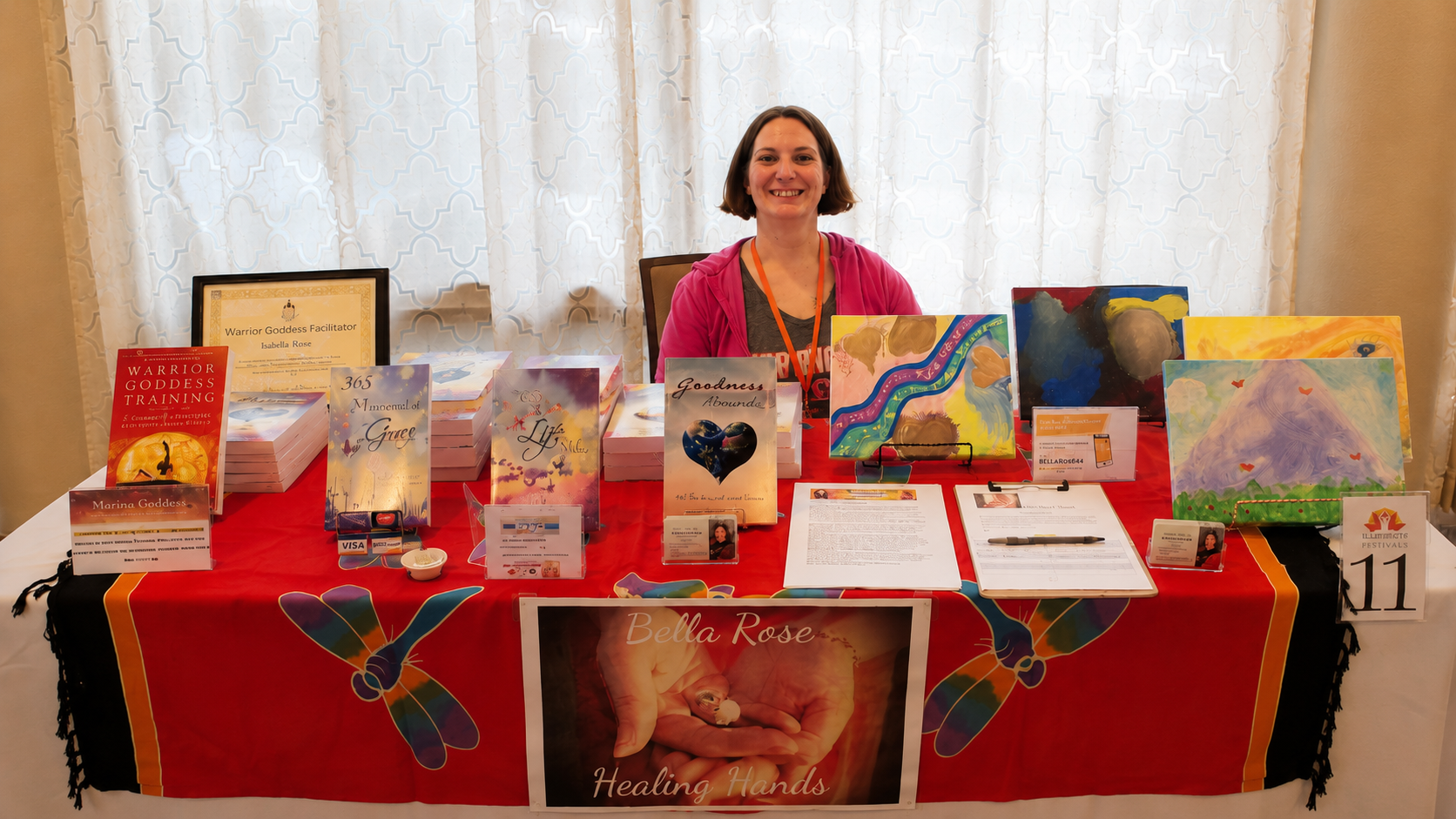 Woman sitting at a table displaying various colorful books and artwork related to healing, spirituality, and goddess training at an indoor event.