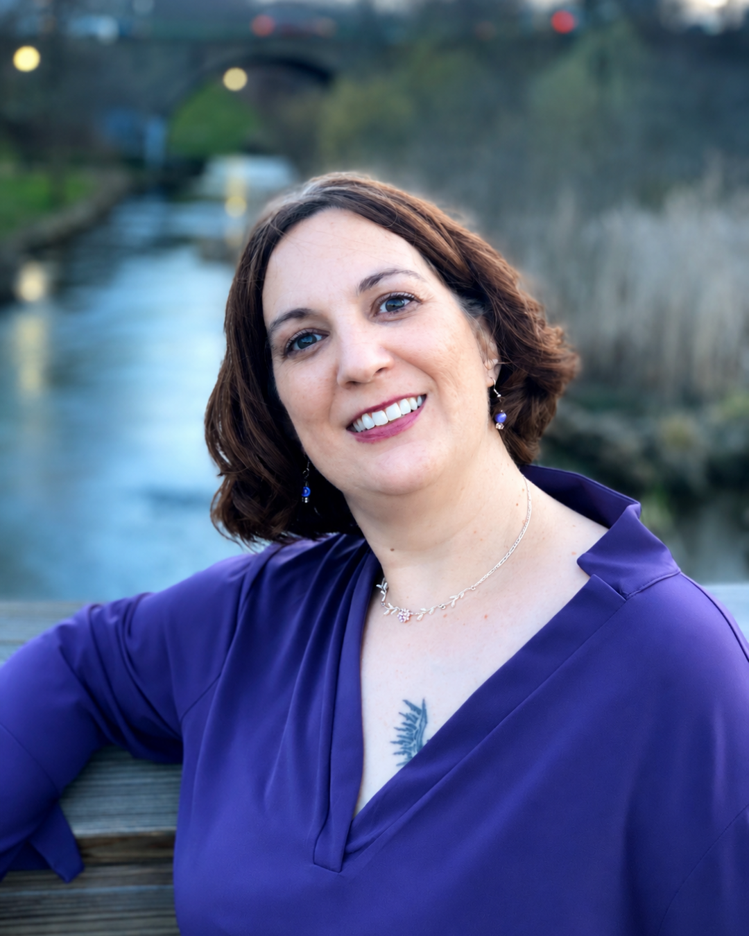 A woman with short brown hair and blue eyes smiling outdoors, wearing a purple top, jewelry, and sitting near a body of water with a bridge in the background.
