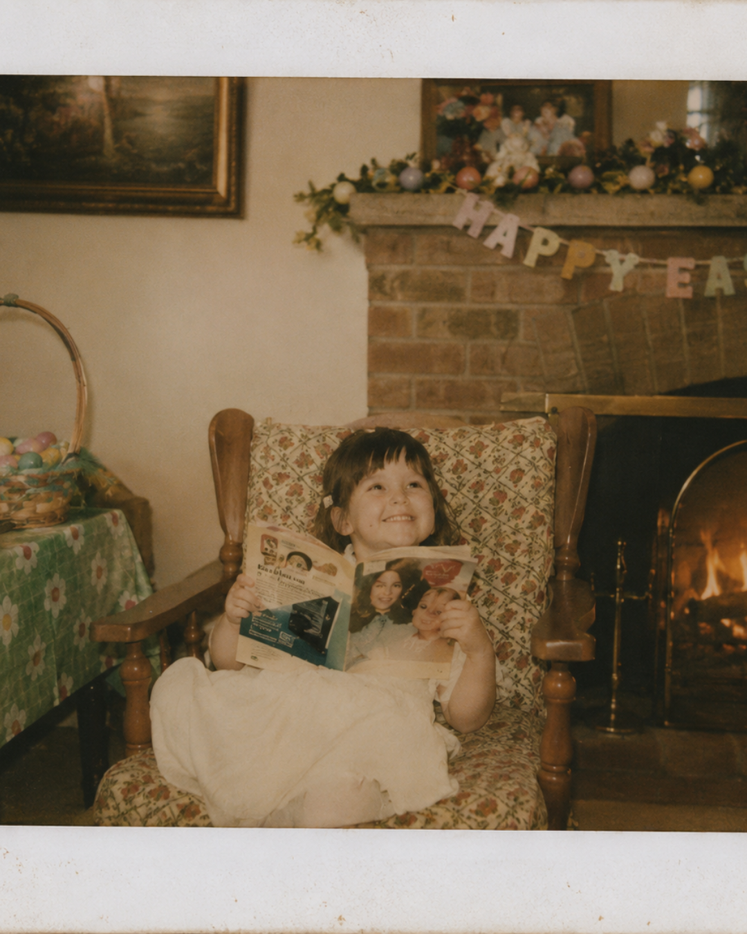 Young girl sitting in a vintage armchair, smiling and holding a magazine, with a fireplace decorated for Easter in the background.