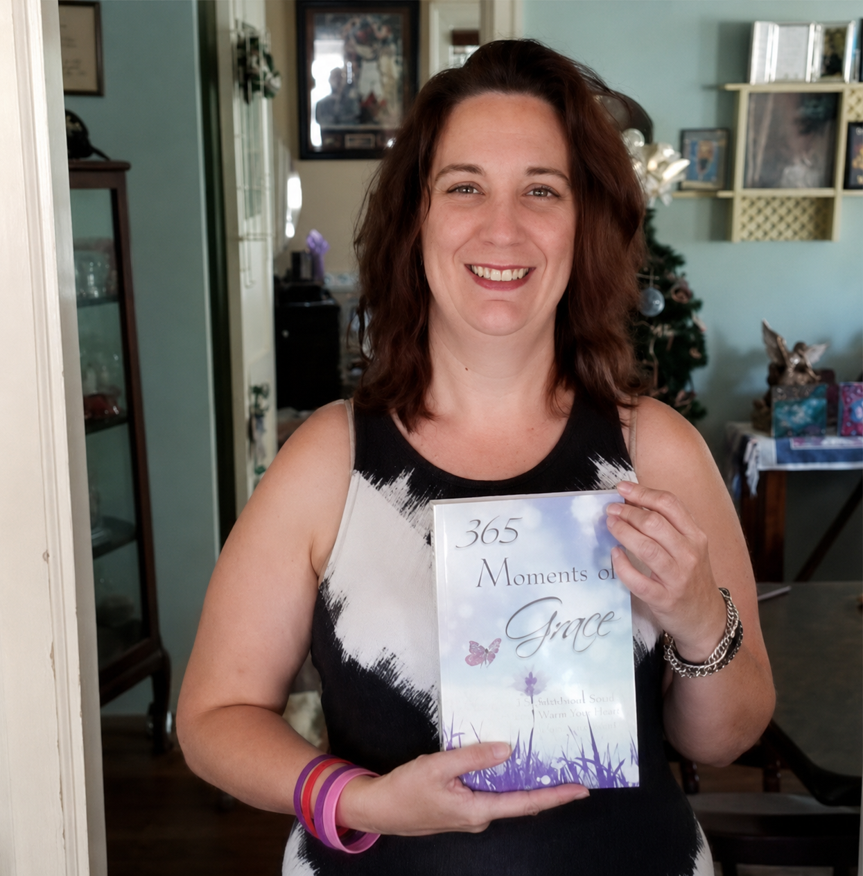 A woman with brown hair smiling and holding a book titled '365 Moments of Grace' in a decorated living room with a Christmas tree, gifts, and a table in the background.