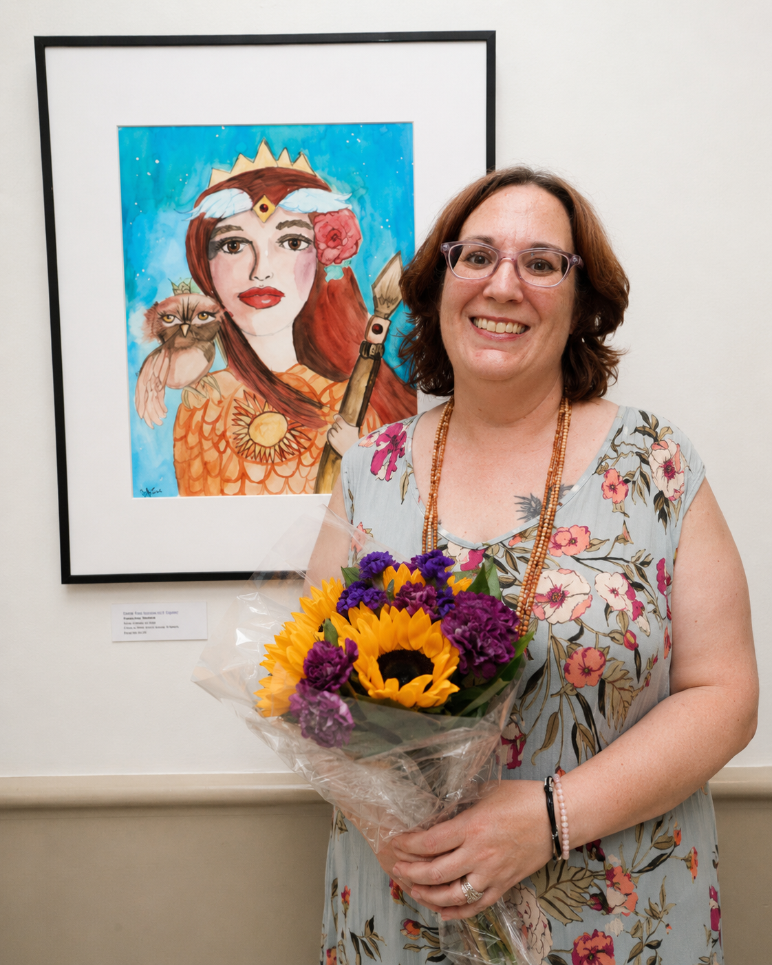 A woman with glasses and wearing a floral dress holds a bouquet of sunflowers and purple flowers, standing in front of a framed colorful illustration of a woman with a crown, a feathered headdress, and an owl.