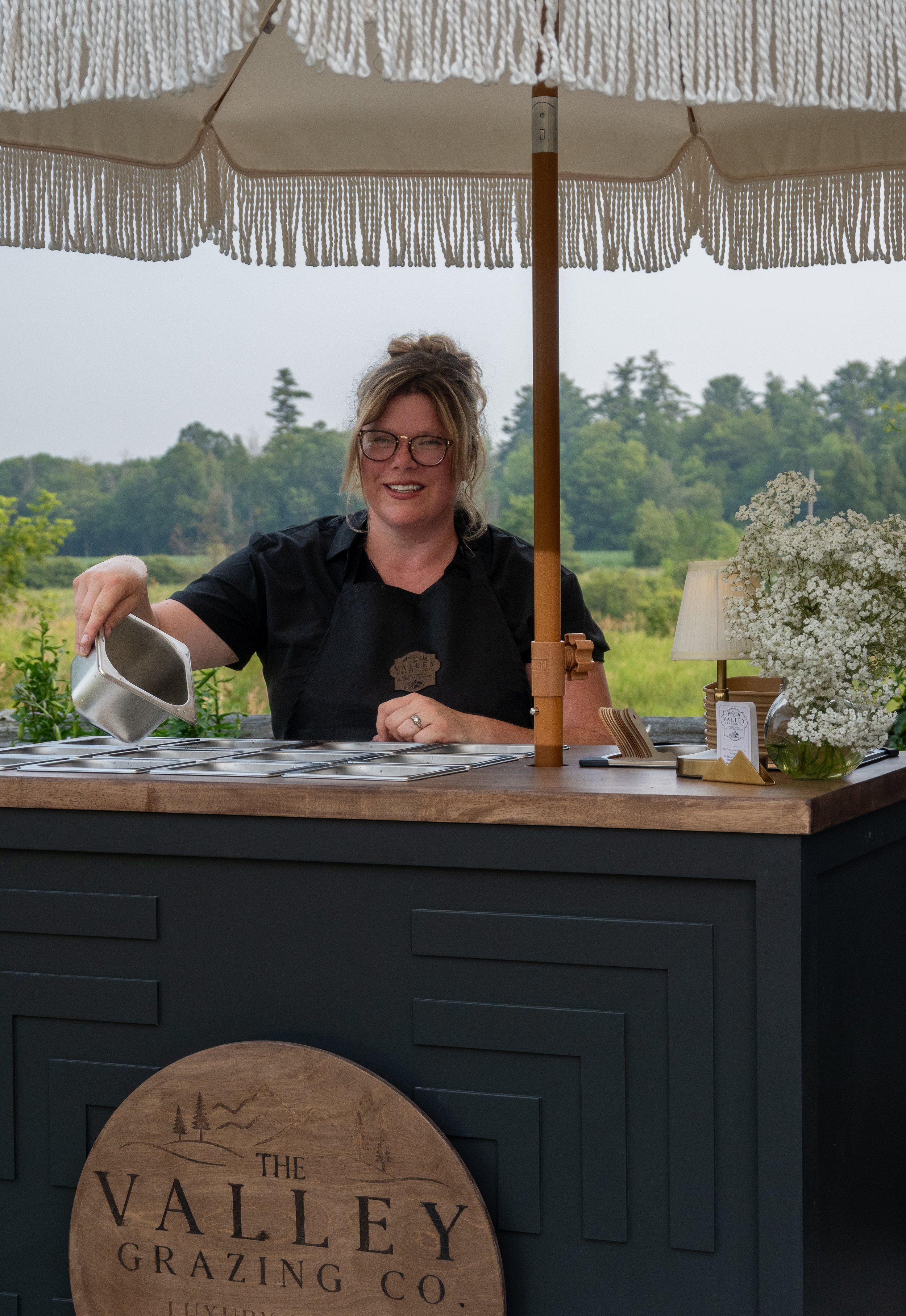 A woman smiling at an outdoor table with an umbrella, flowers, and greenery in the background. She is pouring a beverage and wearing glasses and a black apron. The table has a sign that reads "The Valley Grazing Co.".