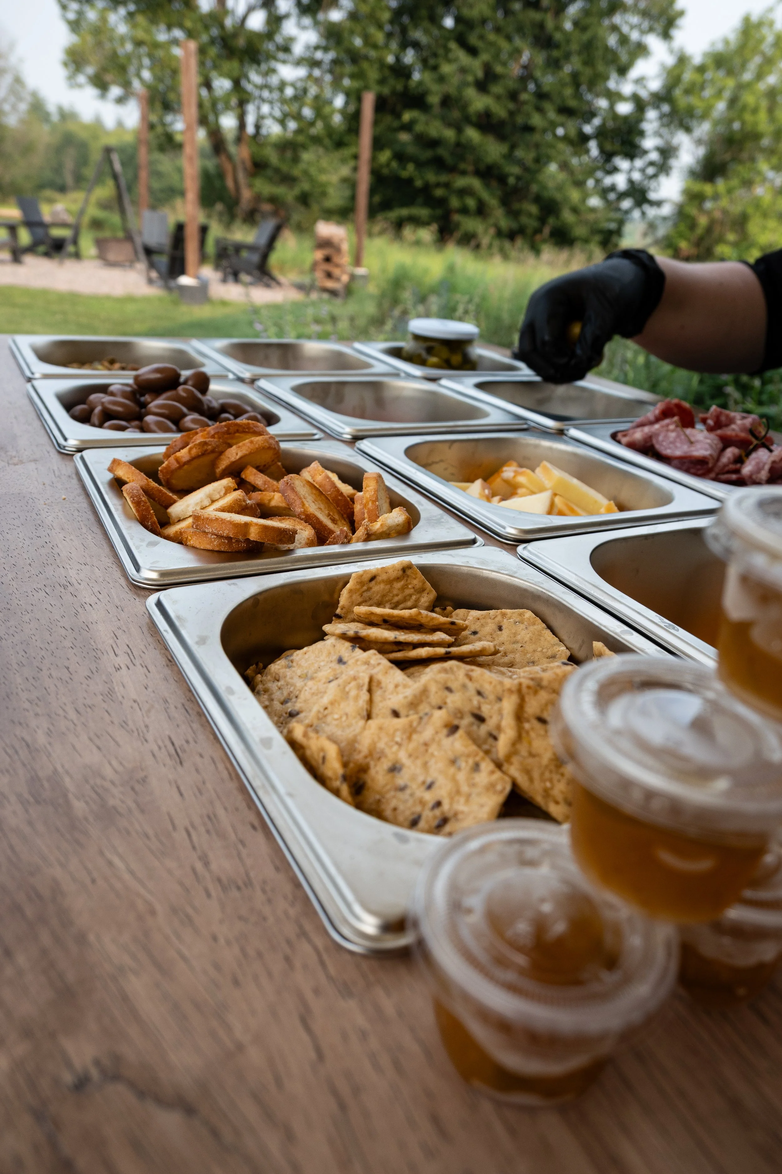 An outdoor buffet setup with various food containers on a wooden table, including crackers, chocolates, cheeses, cold cuts, and honey jars. A person's hand, wearing a black glove, is reaching for a jar of pickles. In the background, there is a grassy