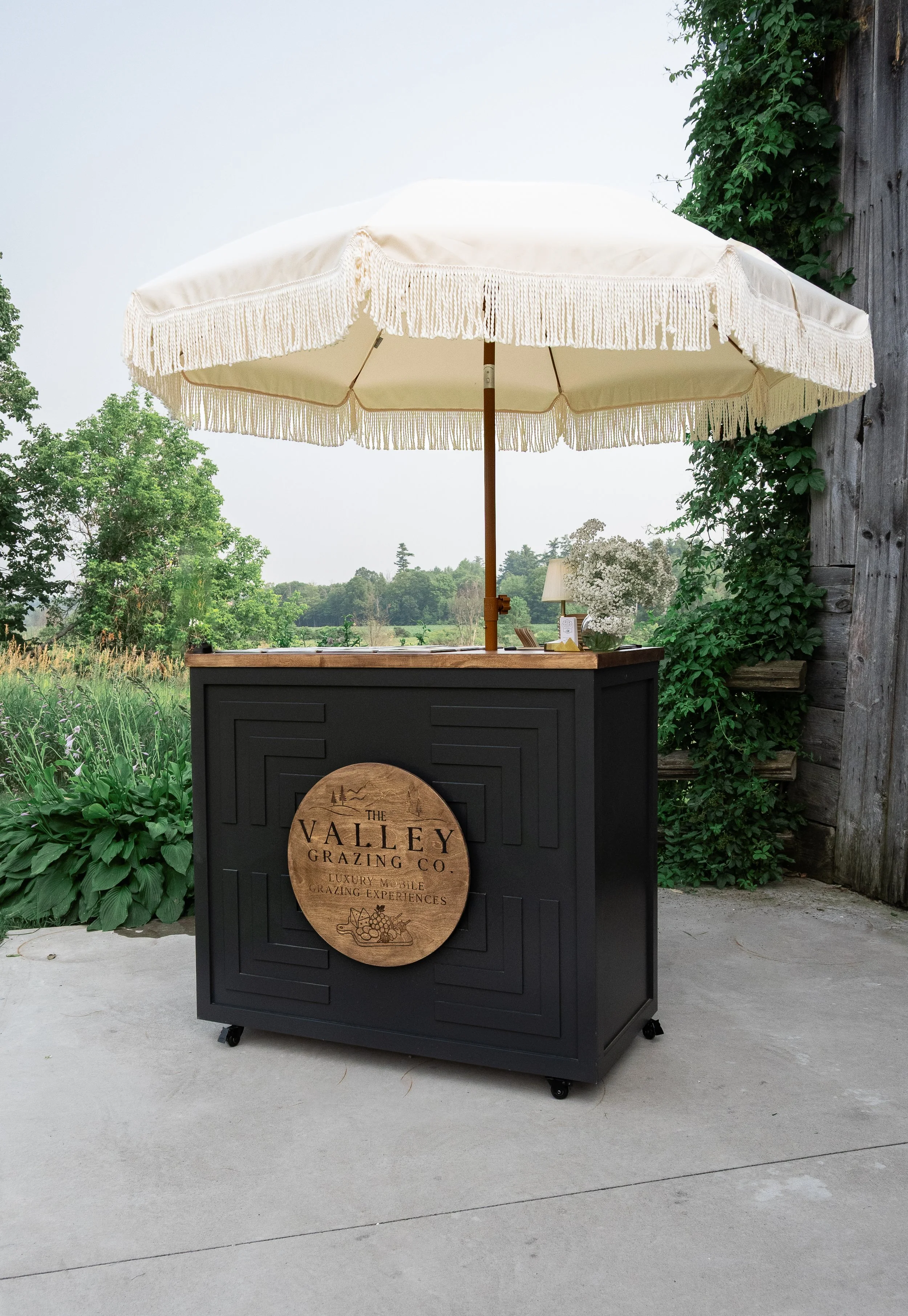 A black mobile grazing station with a wooden sign that says 'The Valley Grazing Co.' and 'Luxury Mobile Grazing Experiences.' It features a large cream-colored parasol with fringe, standing outdoors on a concrete surface with greenery and trees in th