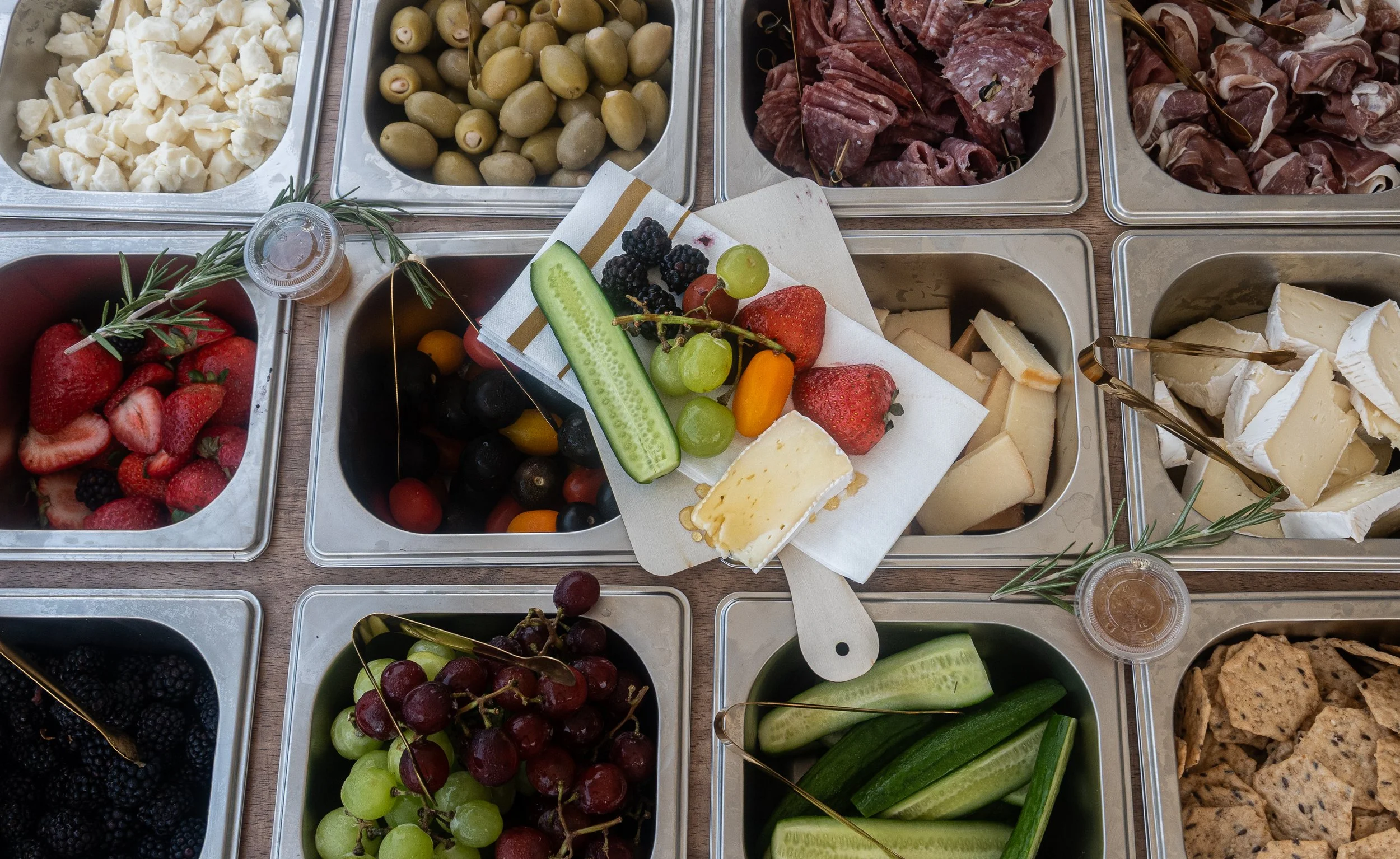 Assorted food items in metal containers including cheese, green olives, pink olives, meats, strawberries, blackberries, grapes, cucumbers, and crackers.