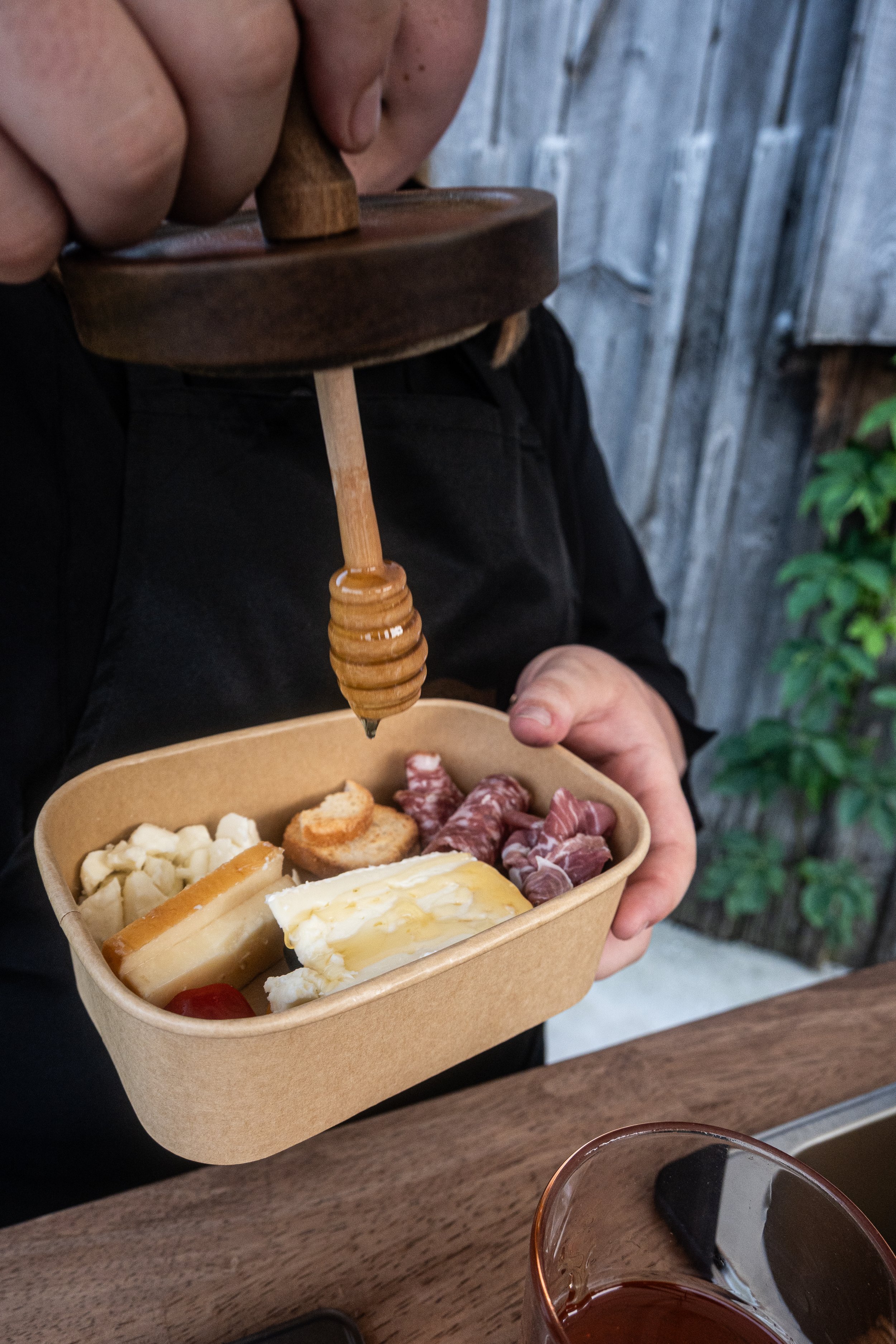 A person holding a biodegradable takeout container filled with cheese, grapes, crackers, and cured meats, using a wooden honey dipper to serve honey over the cheese; a glass of red wine on a wooden table, with a rustic wooden wall background and some