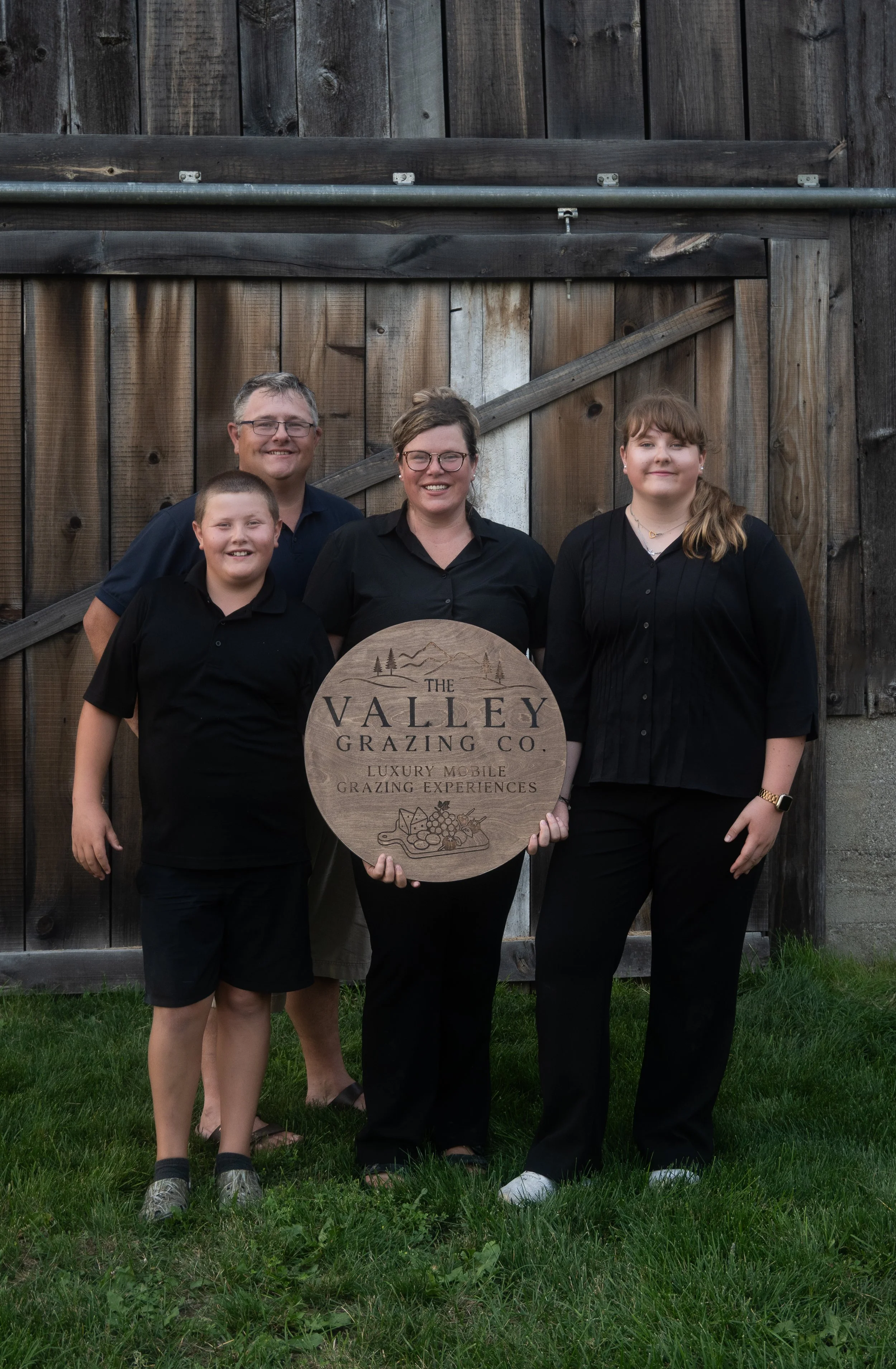 Four people, two adults and two children, standing outside on grass in front of a wooden barn, holding a wooden sign that reads "The Valley Grazing Co., Luxury Mobile Grazing Experiences."