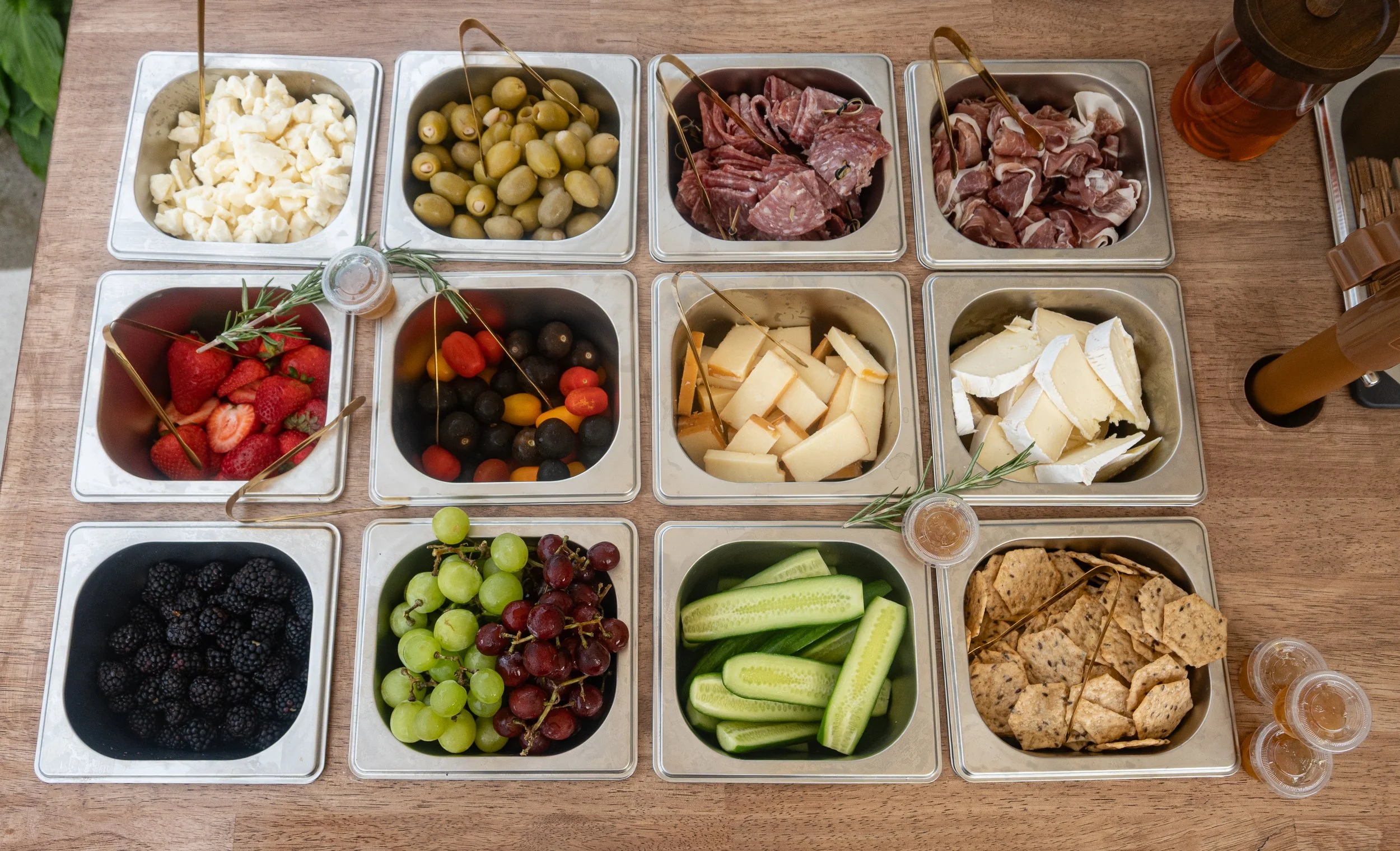 A selection of cheese, meats, fruits, and vegetables in metal containers arranged on a wooden table, with small condiment cups.