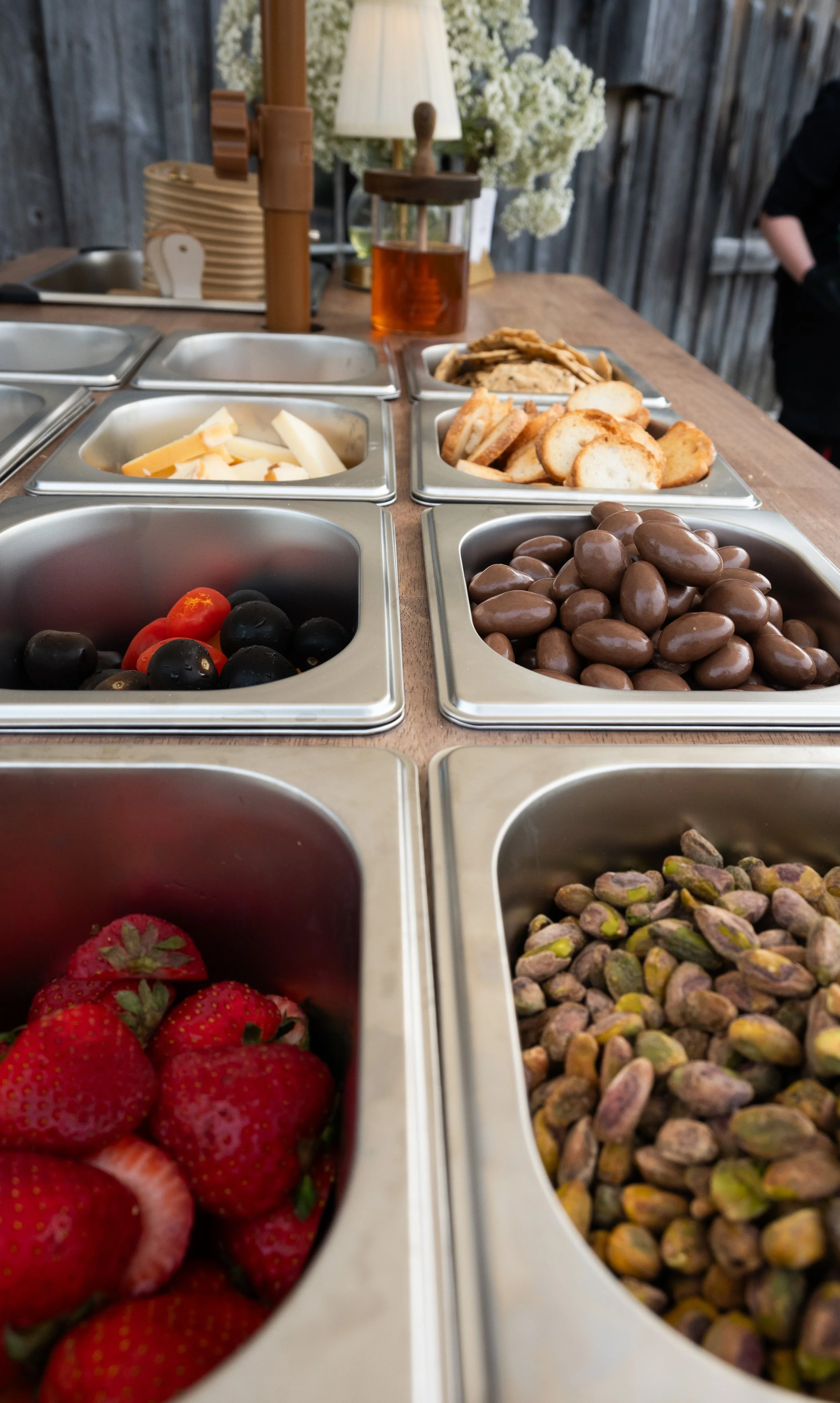 View of various toppings in metal containers at a self-serve topping bar, including strawberries, blueberries, chocolate-covered nuts, pistachios, and other nuts.