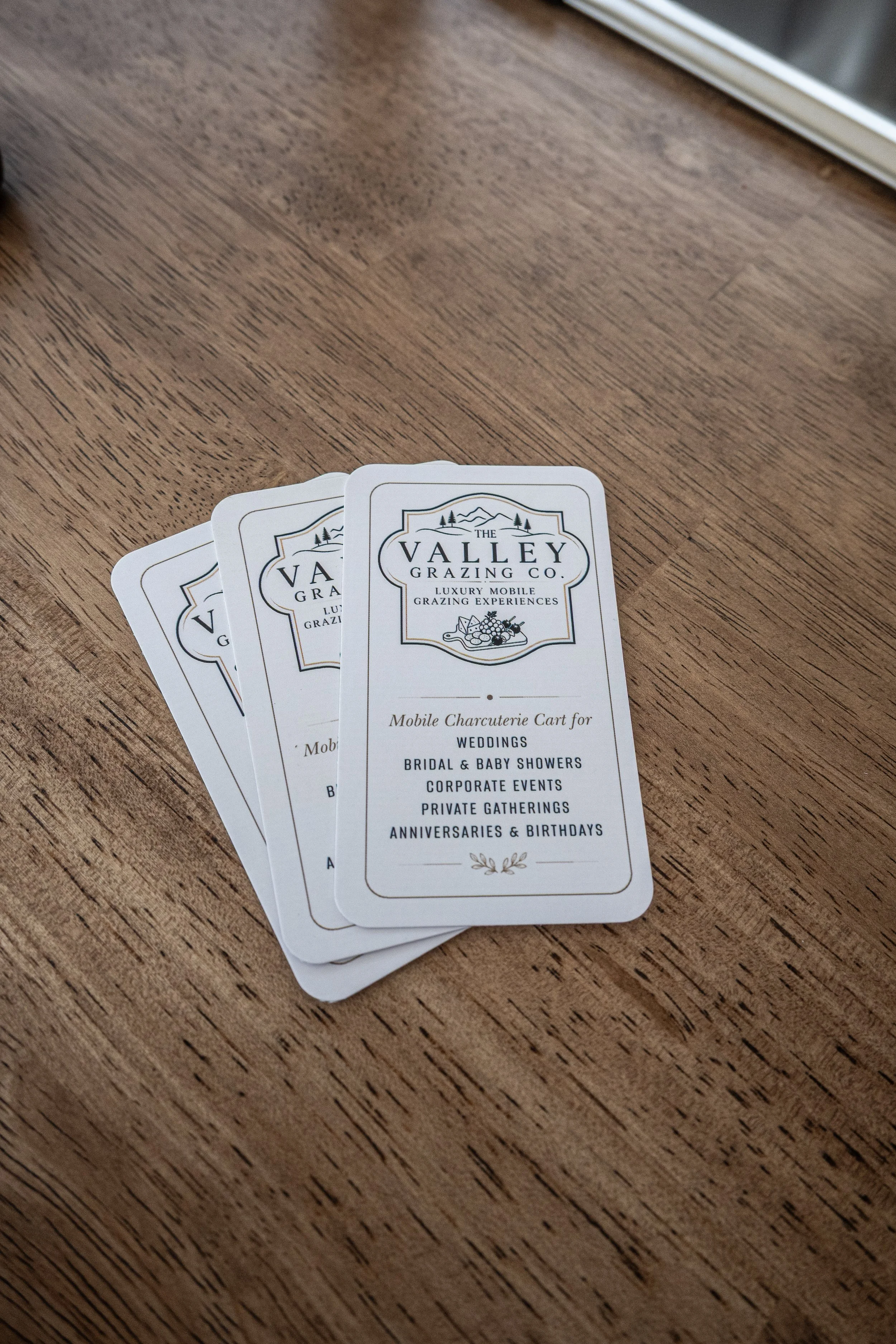 Business cards for The Valley Grazing Co. are fanned out on a wooden surface. The cards mention mobile charcuterie cart services for weddings, bridal and baby showers, corporate events, private gatherings, and birthdays.