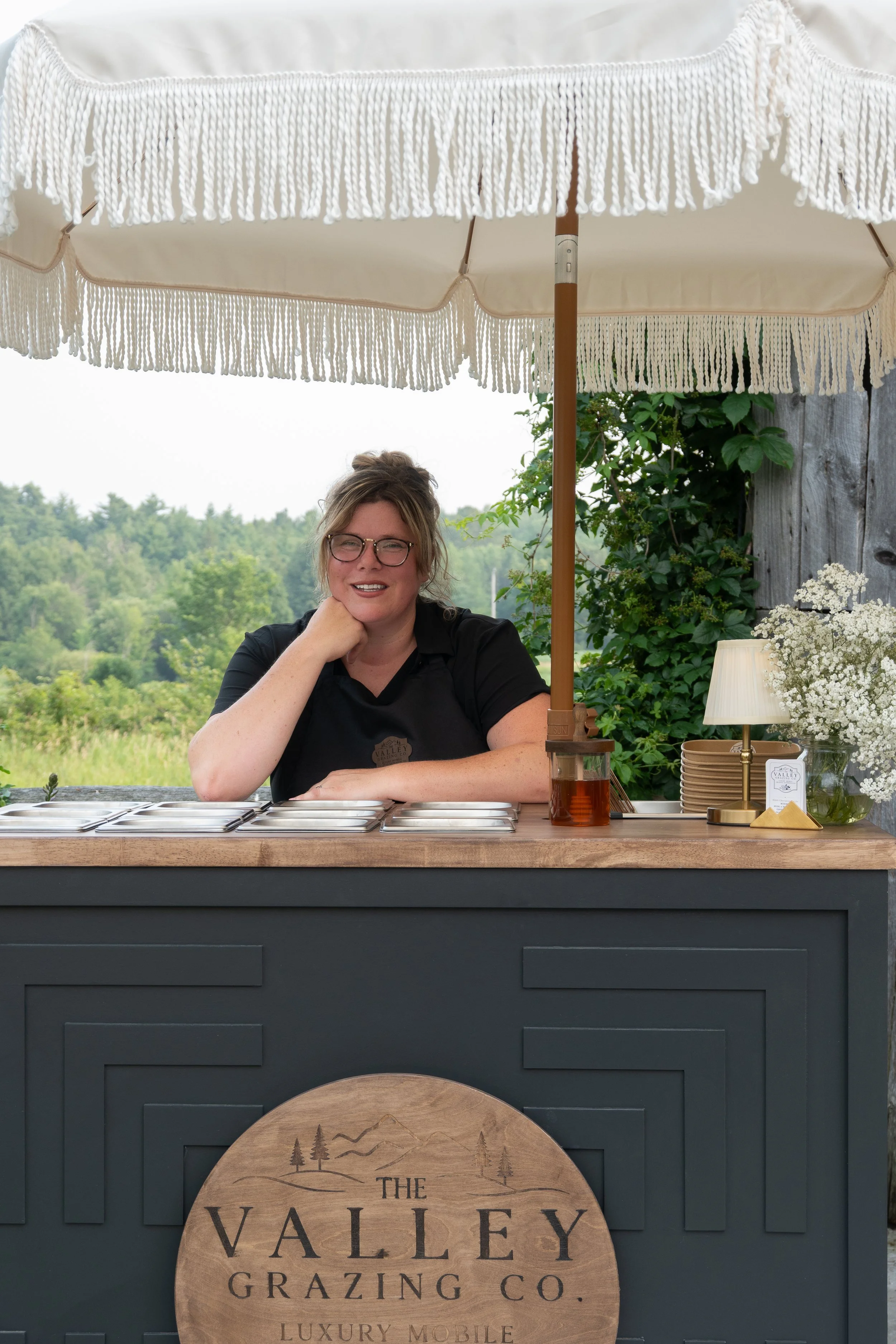 A woman sitting at an outdoor booth with a large sun umbrella, greenery in the background, and a wooden sign that reads "The Valley Grazing Co."