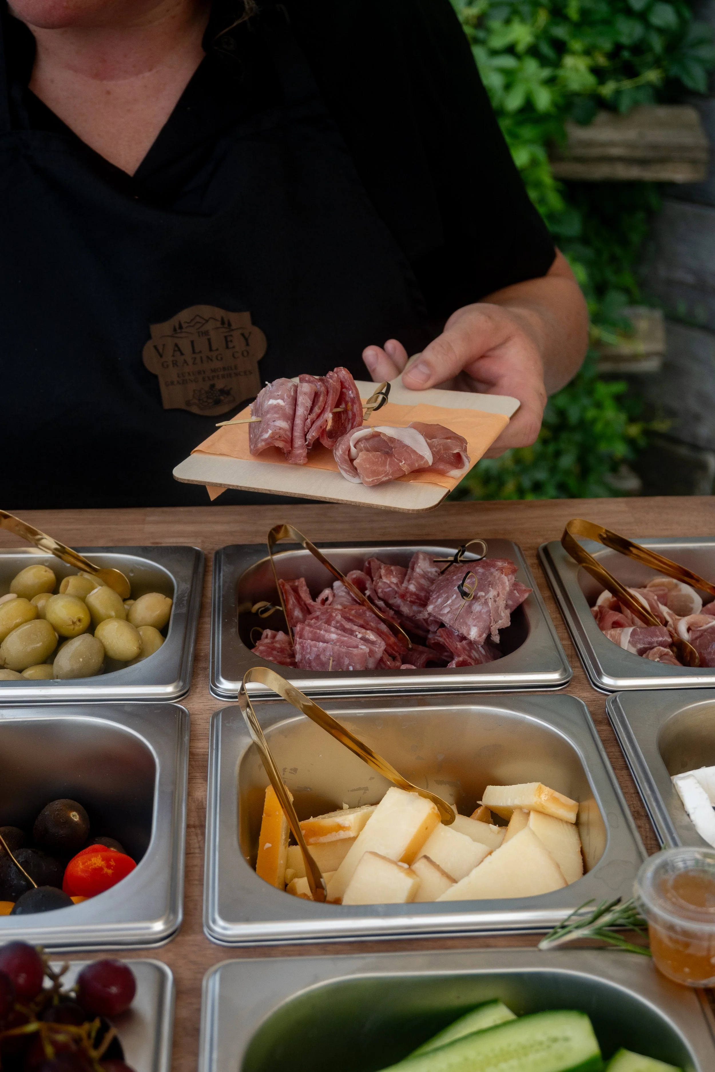 A person in black shirt with a Valley Grazing Co pin holding a small wooden platter with sliced cured meats, situated at a buffet with various cheeses, fruits, and vegetables in metal containers.