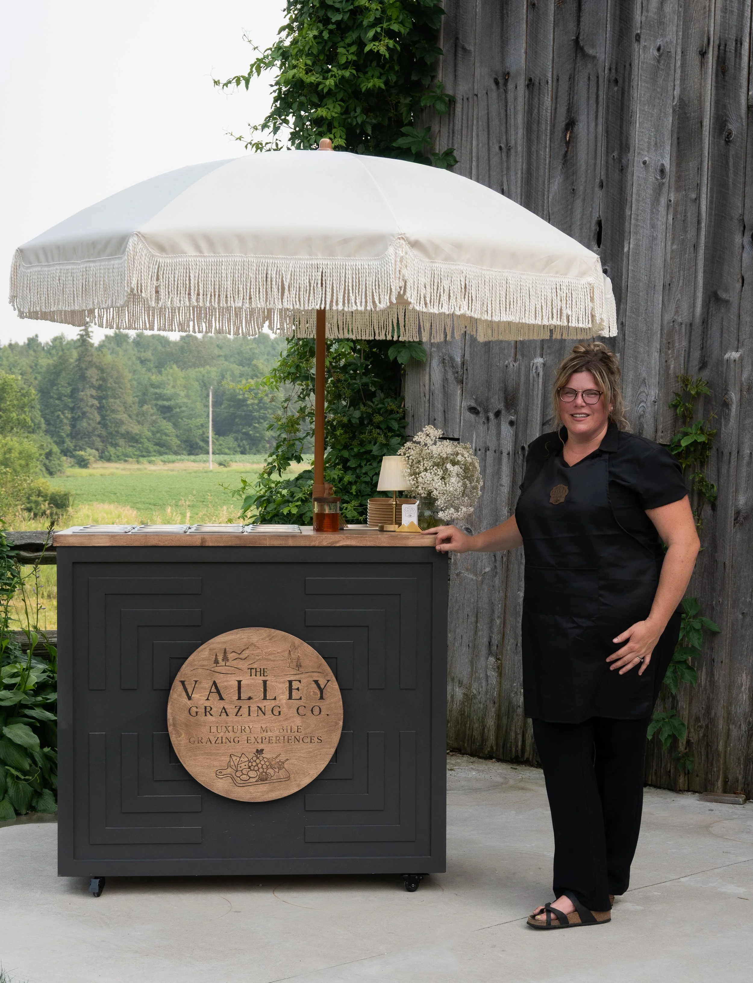 A woman in black scrubs standing next to a black mobile grazing cart with a wooden sign reading 'The Valley Grazing Co. Luxury Mobile Grazing Experiences,' in front of a rustic wooden wall, under a large white umbrella, with a green field and trees i