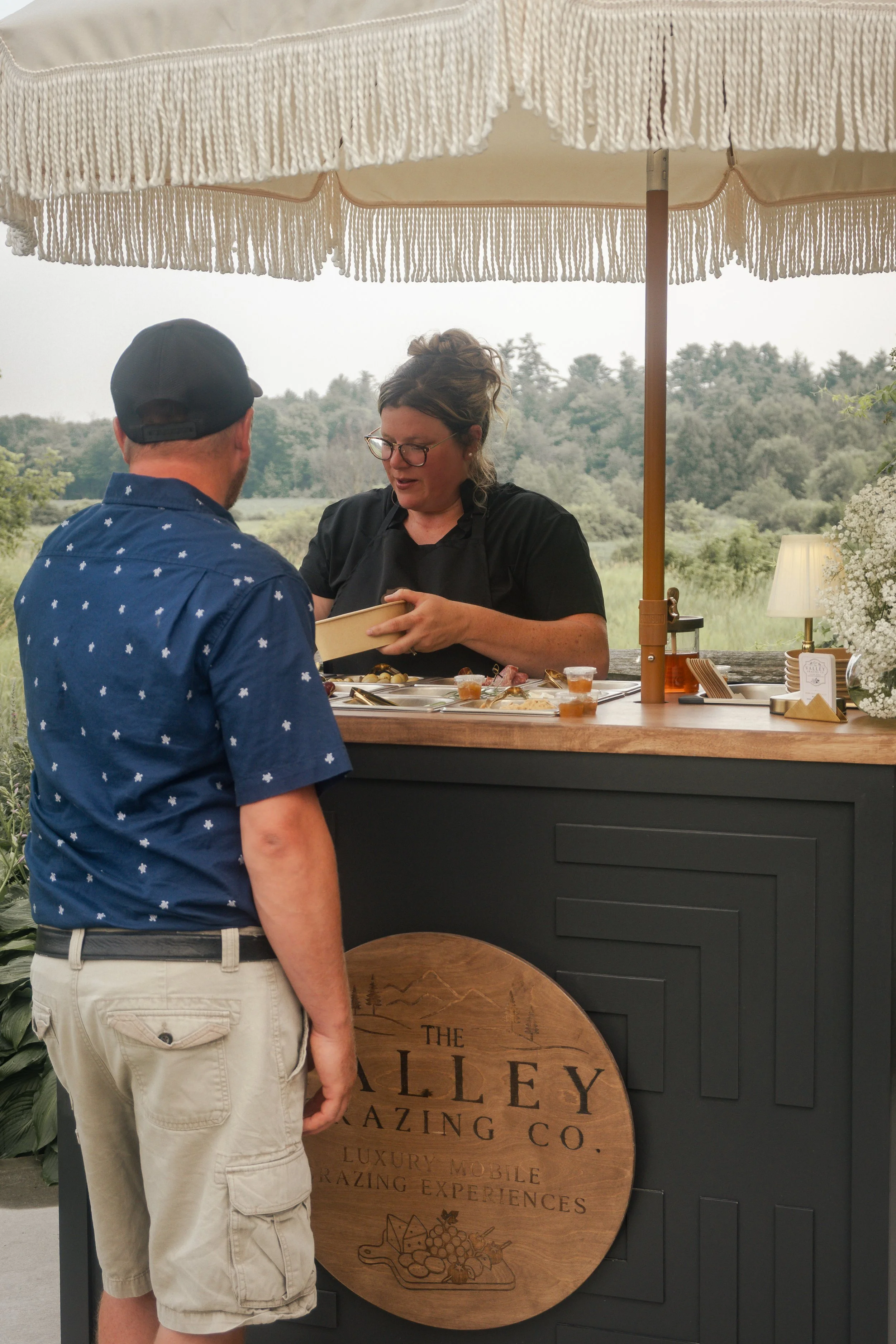 A man in a blue shirt and beige shorts stands in front of a food stand while a woman behind the counter speaks to him. The stand has a large round wooden sign with text and a fruit illustration, a white parasol, and is set outdoors with trees in the background.