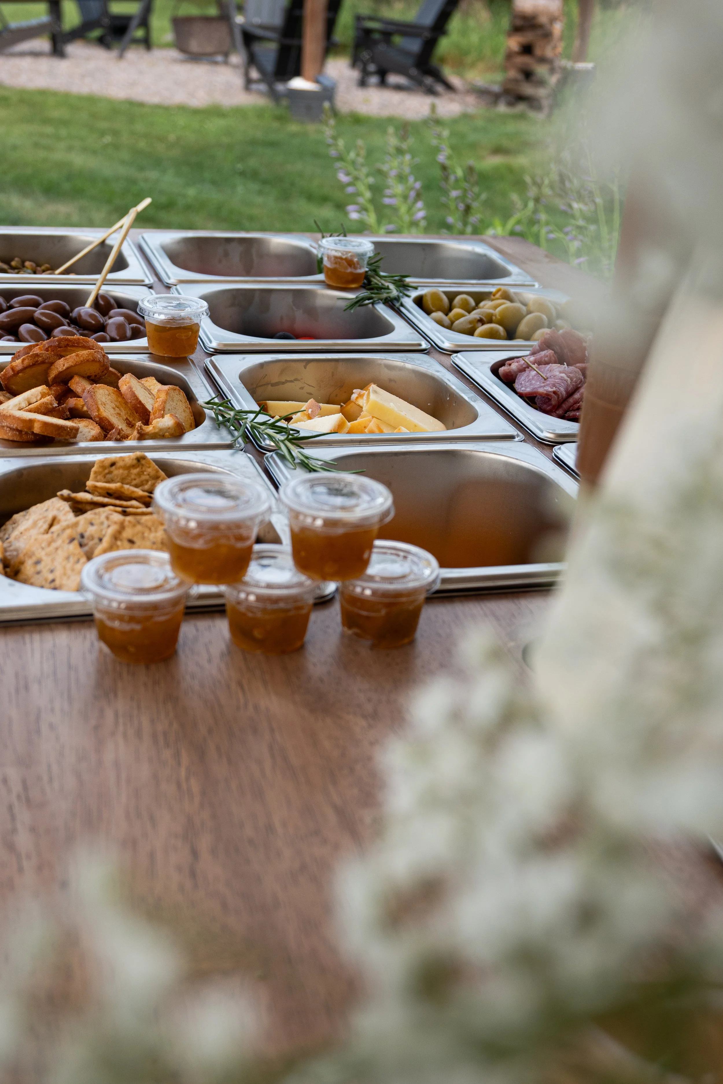 Outdoor buffet setup with various appetizers and condiments, including cheese, olives, pickles, and jars of honey, on a wooden table against a grassy background.