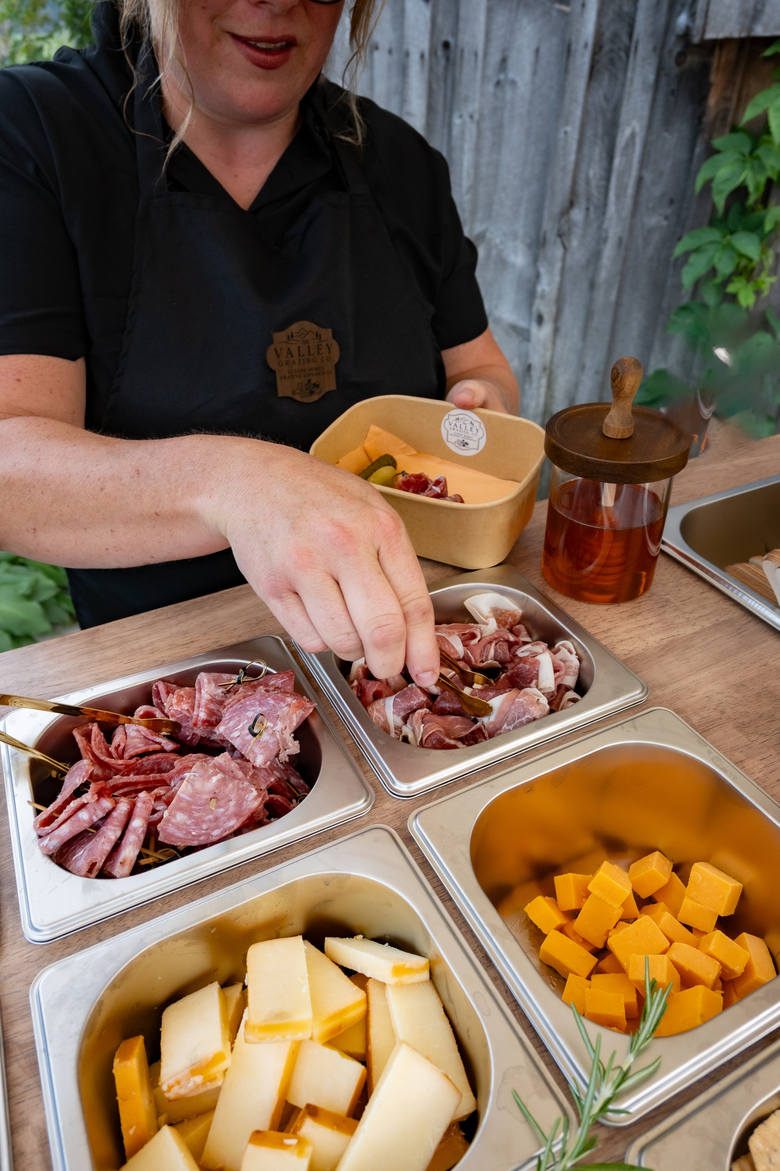 Person serving a platter of sliced cheese on a wooden table with various cheeses and sliced meats in metal trays.