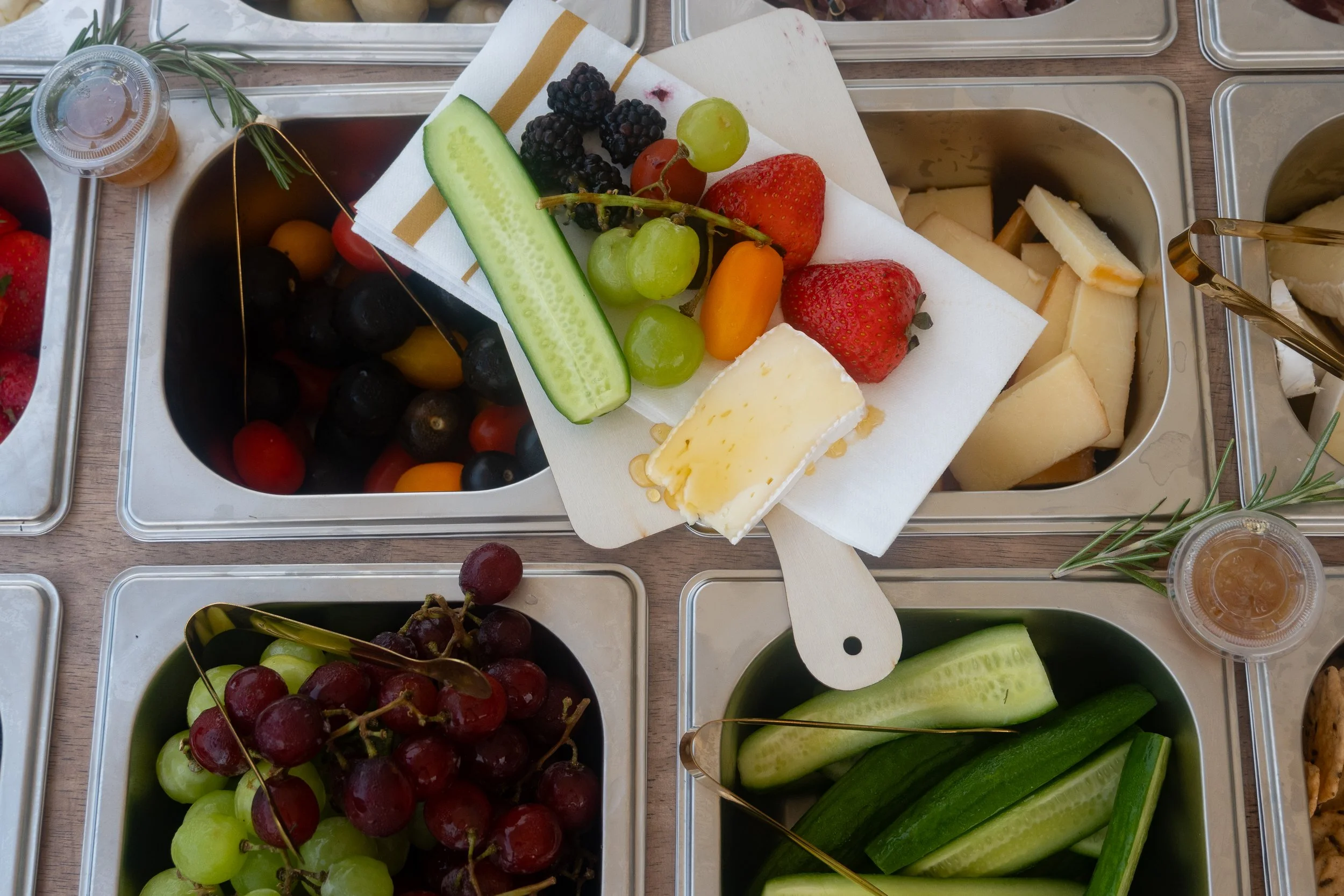 A tray of fresh fruits including strawberries, green grapes, blackberries, a sliced cucumber, and a cherry tomato, with blocks of cheese in a stainless steel container nearby.