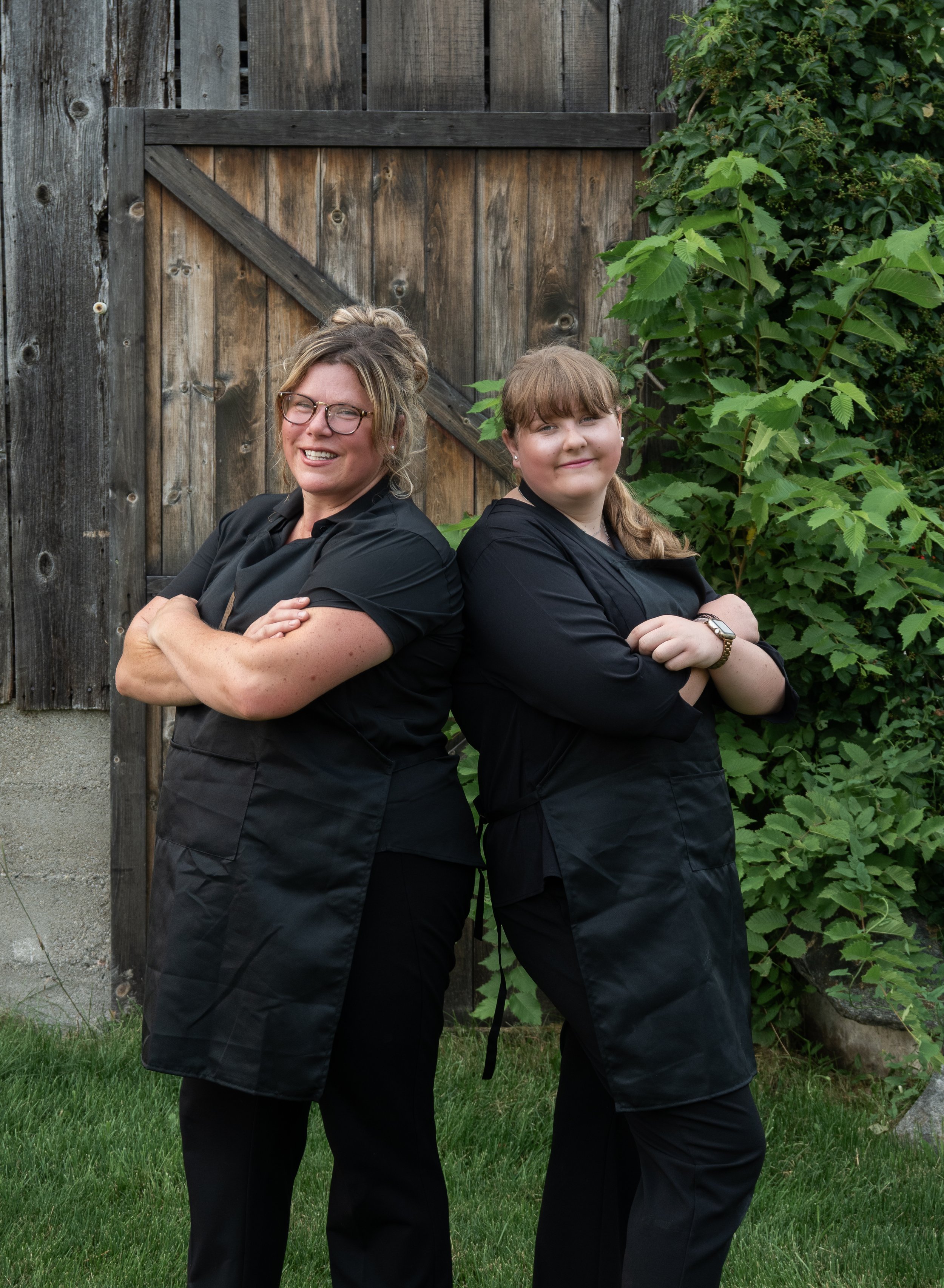 Two women with their arms crossed, smiling and standing back to back outdoors in front of a wooden fence and green foliage.