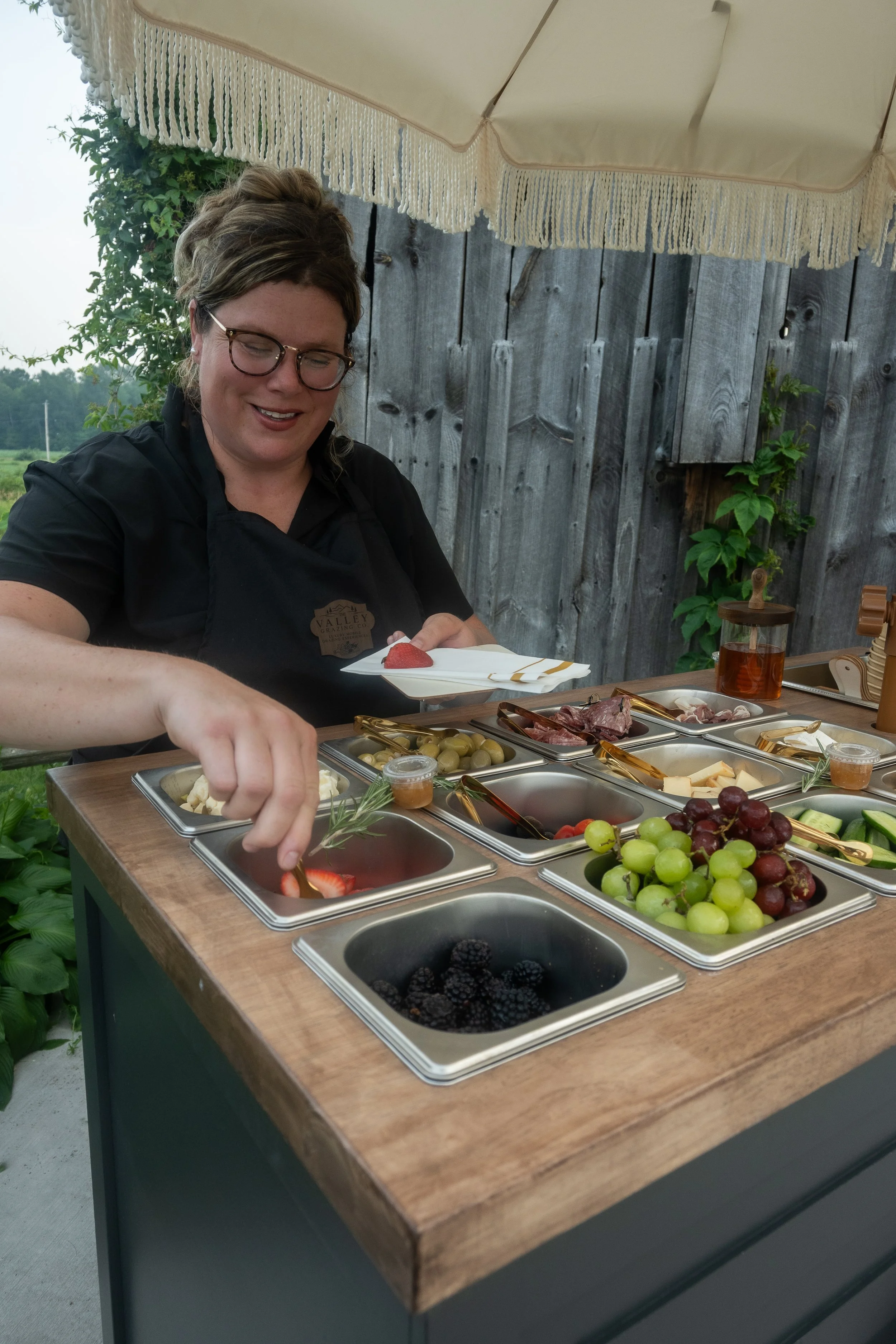 A woman with glasses in a black apron serving herself fruit from a buffet table outdoors, with a gray wooden fence and green plants in the background. The table has stainless steel trays filled with various fruits including strawberries, grapes, blac