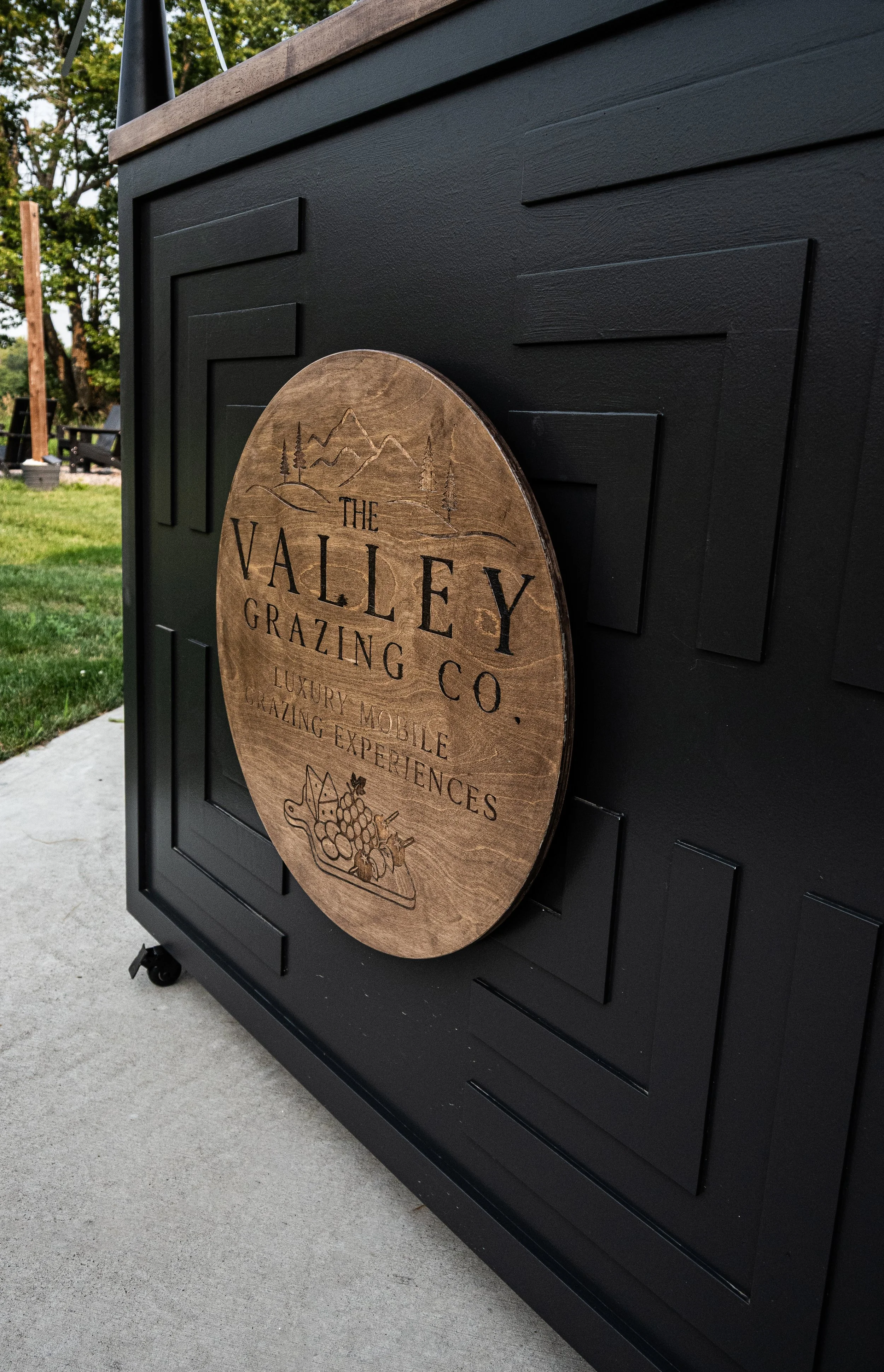 Close-up of a black outdoor cabinet with a round wooden sign attached. The sign reads 'The Valley Grazing Co.' and mentions luxury mobile grazing experiences. The background shows green lawn, trees, and outdoor furniture.