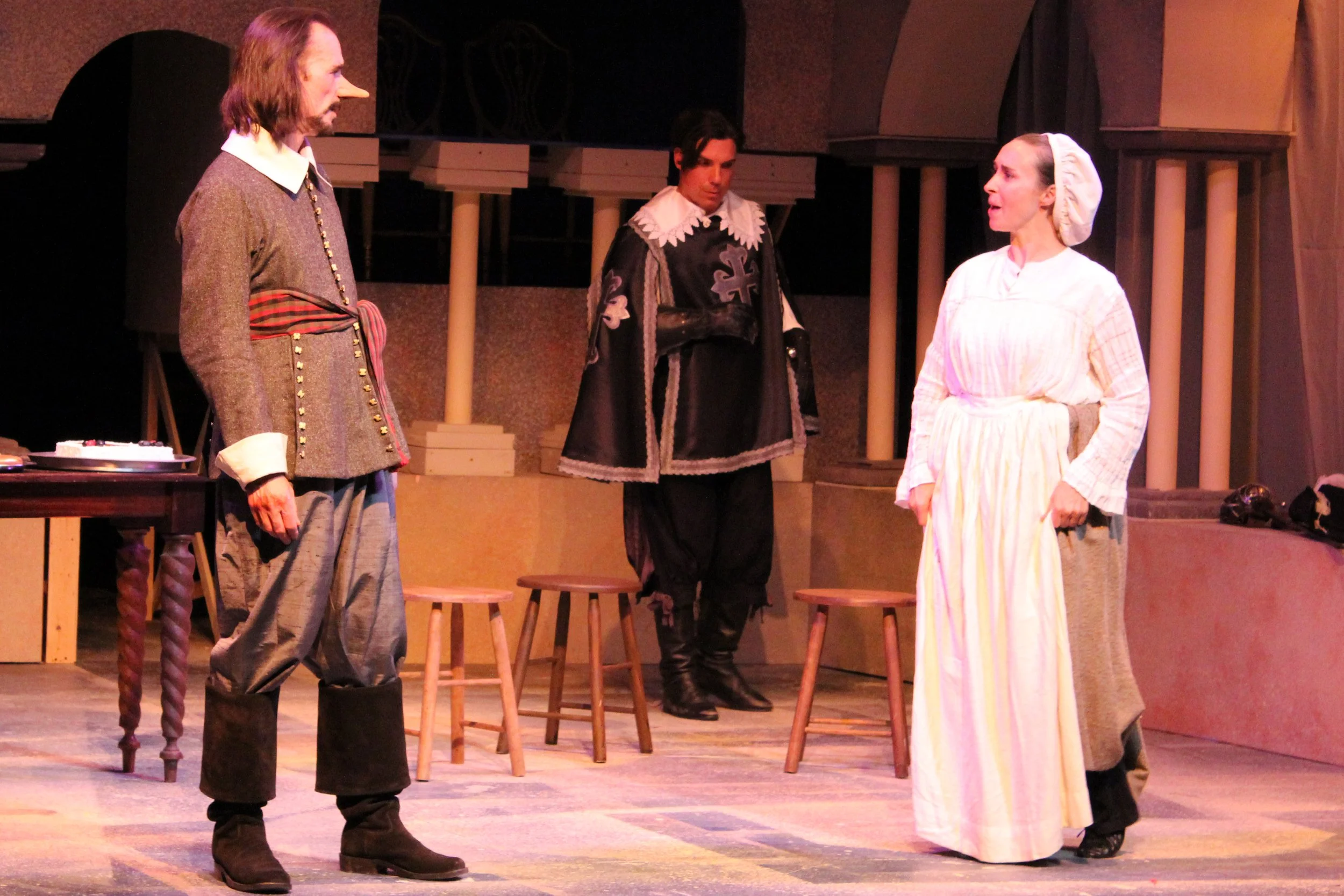 Three actors in costume performing on stage, with one man on the left dressed in historical clothing, a man in the middle dressed as a knight, and a woman on the right dressed as a woman from a historical period, engaged in a theatrical scene.
