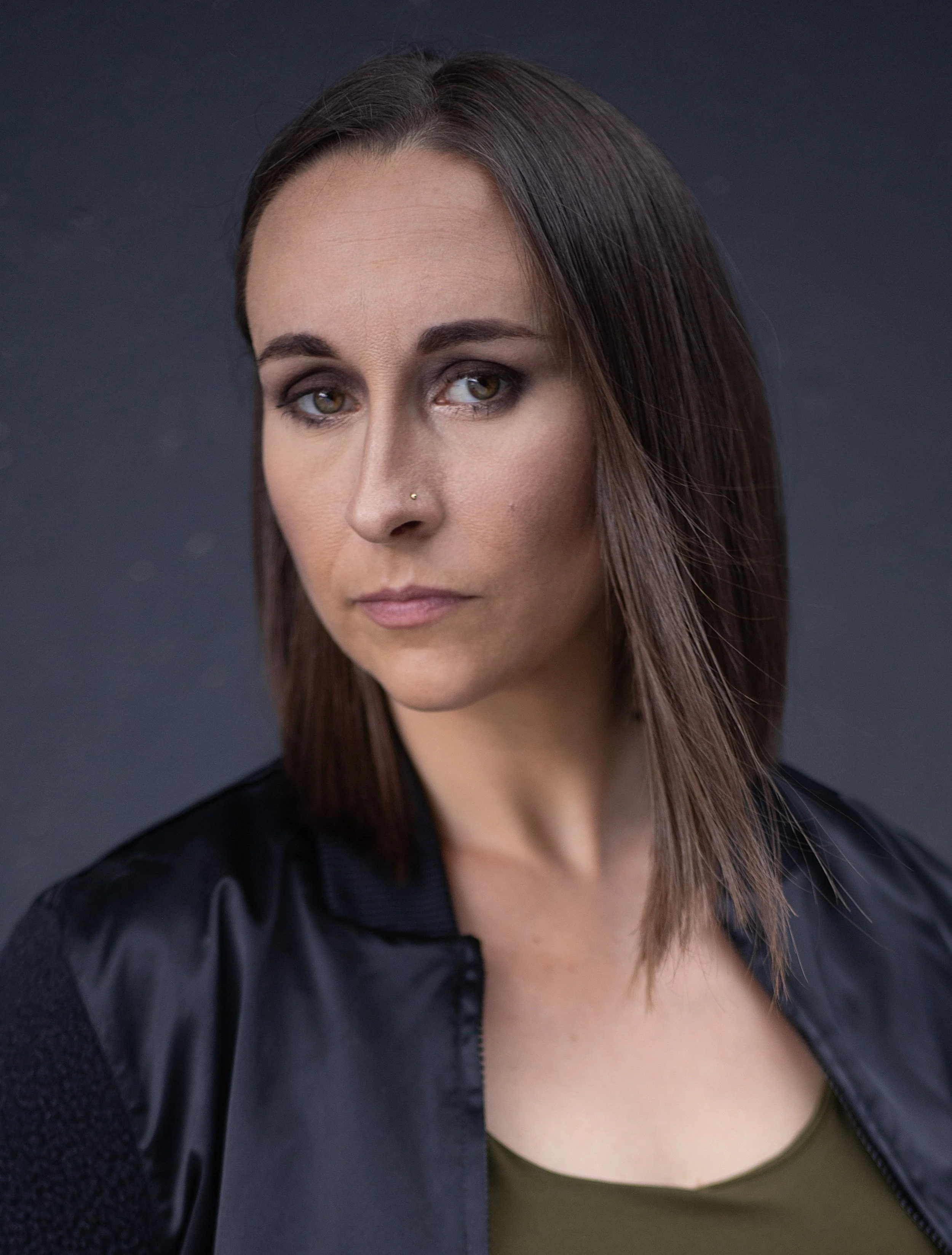 Close-up of a woman with brown hair, wearing makeup including eyeshadow and lipstick, with a nose piercing, wearing a green top and a black jacket, against a dark background.